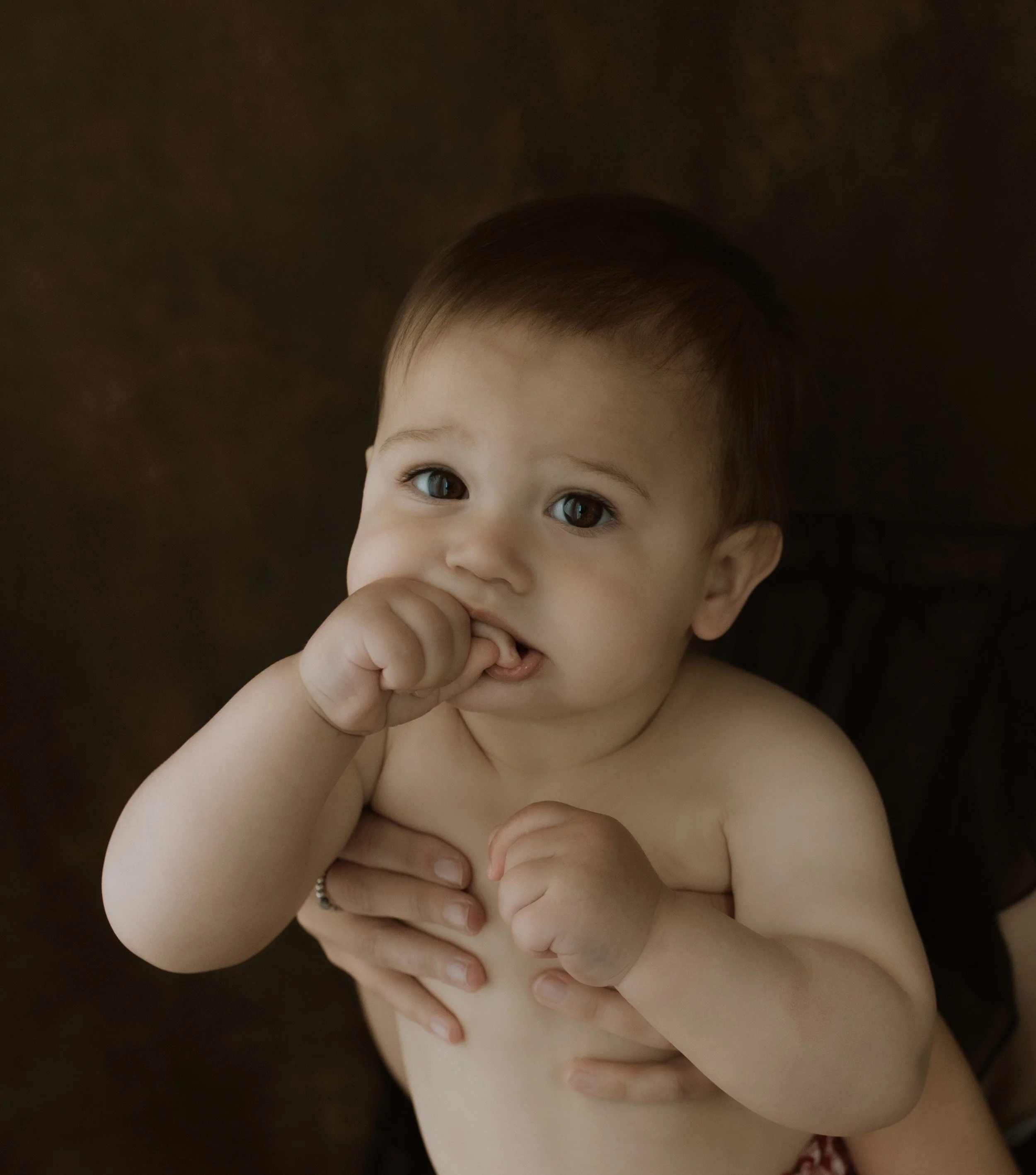 Close-up of a baby with light skin and dark hair, holding their hand near their mouth, against a dark background.