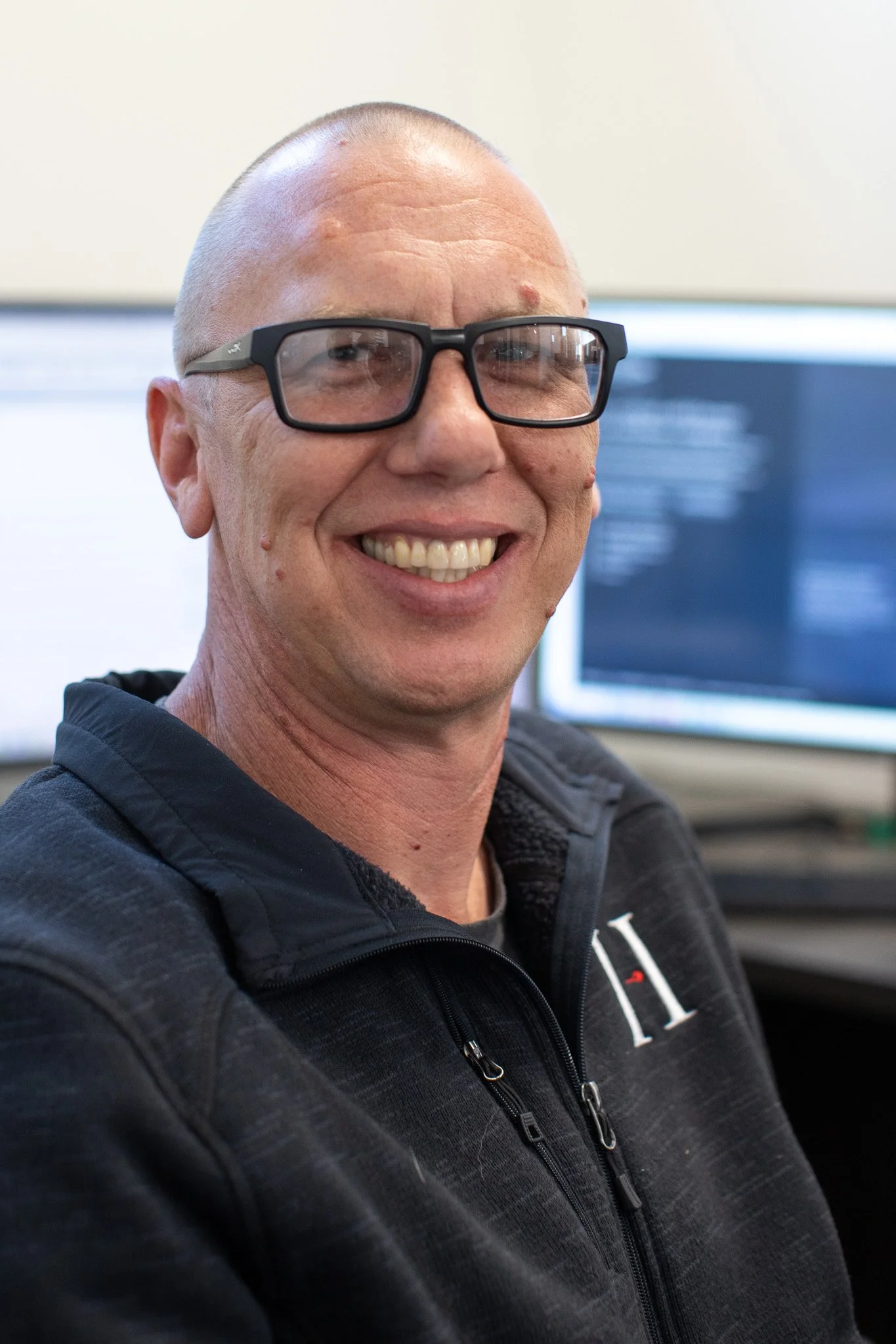 A man with glasses smiling, wearing a black zip-up hoodie, and standing in front of computer monitors in an office setting.