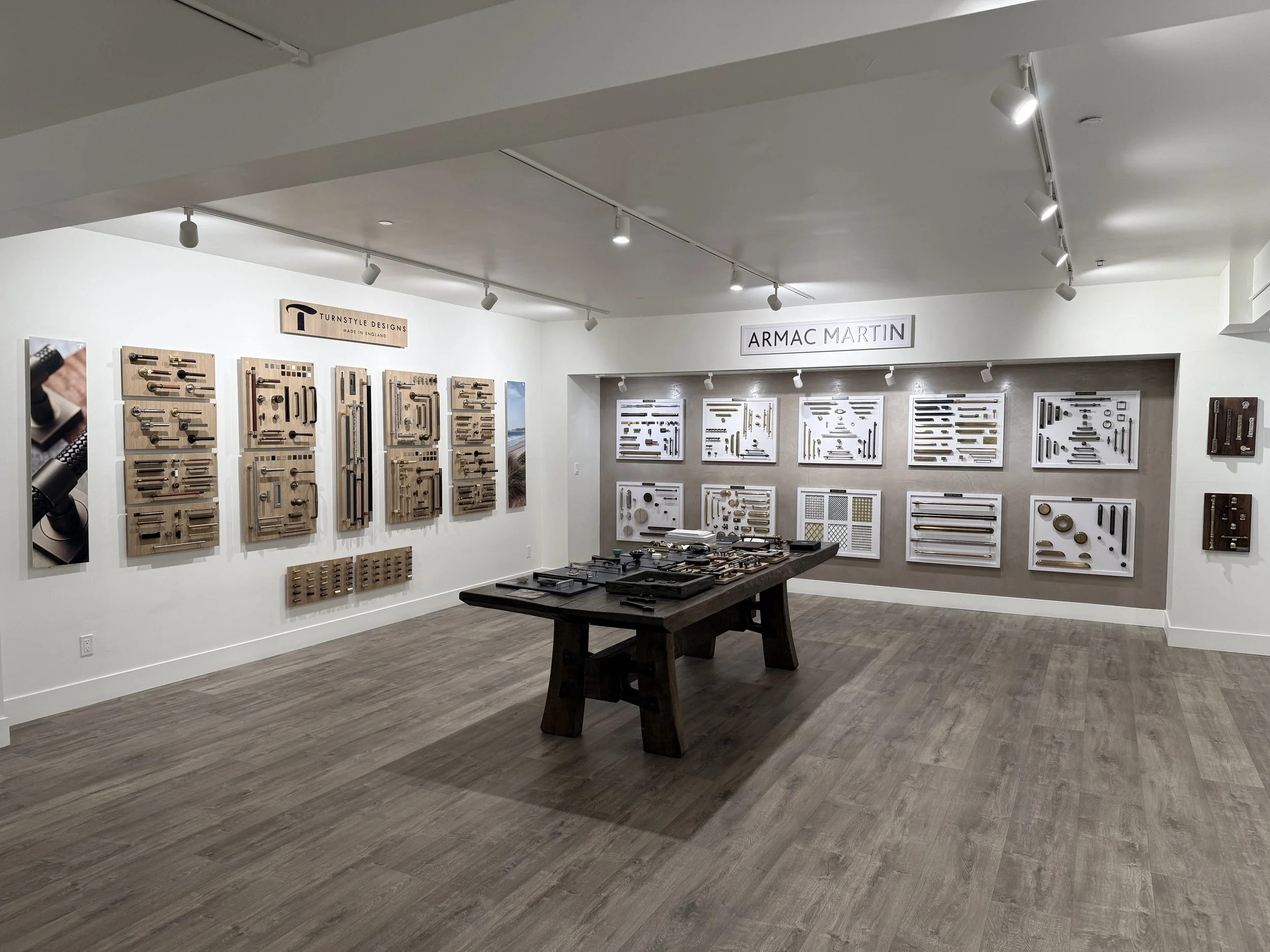 An interior showroom displaying hardware components such as handles, rods, and brackets on white and light beige wall panels, with a dark wooden table in the center, under ceiling spotlights and a sign reading 'ARMA MARTIN'.