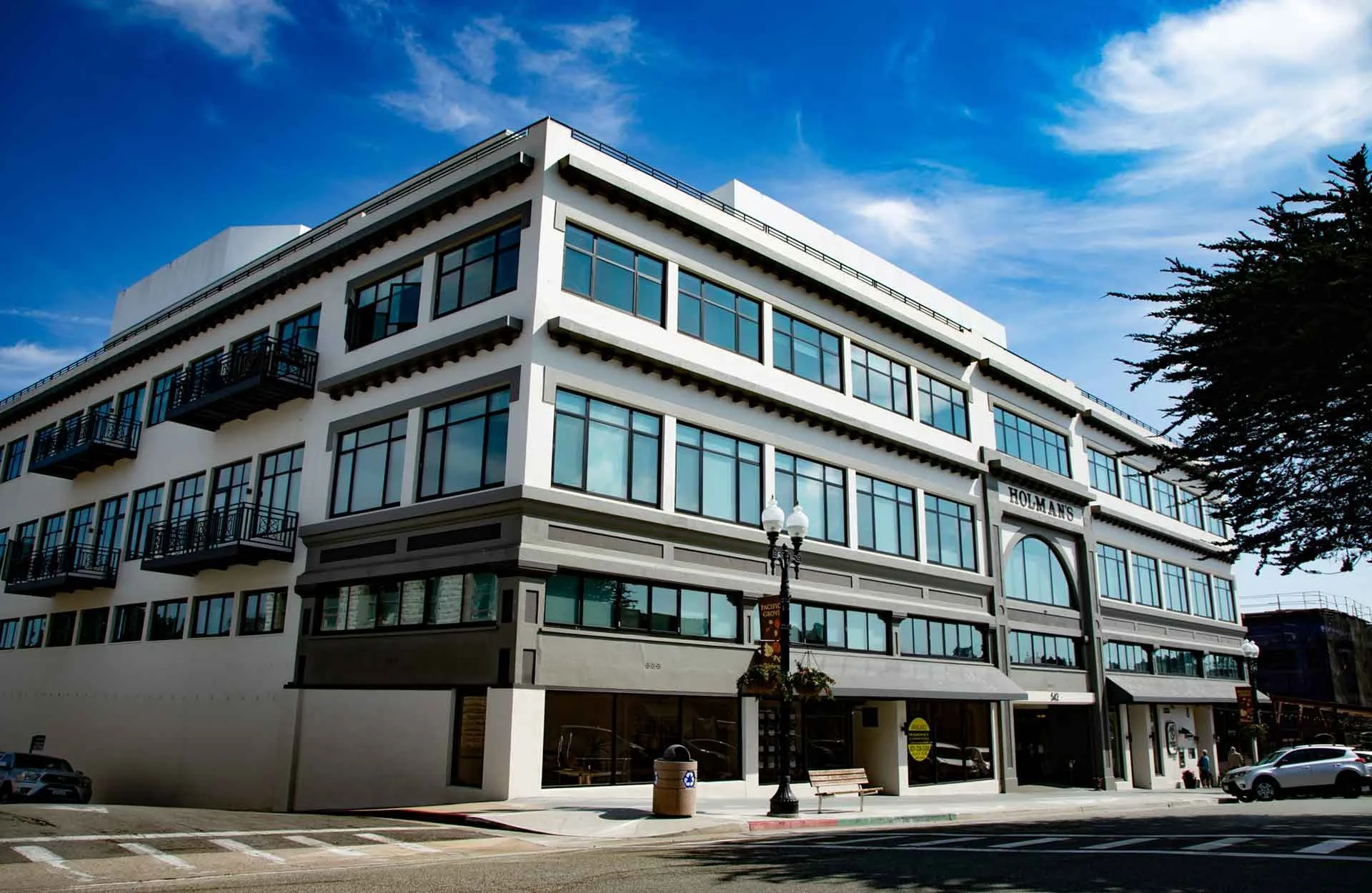 A modern multi-story building with large glass windows, balconies, and a sign that reads 'Holman's' on the front, with a blue sky and clouds above. There are cars parked in front and a streetlamp and bench near the sidewalk.