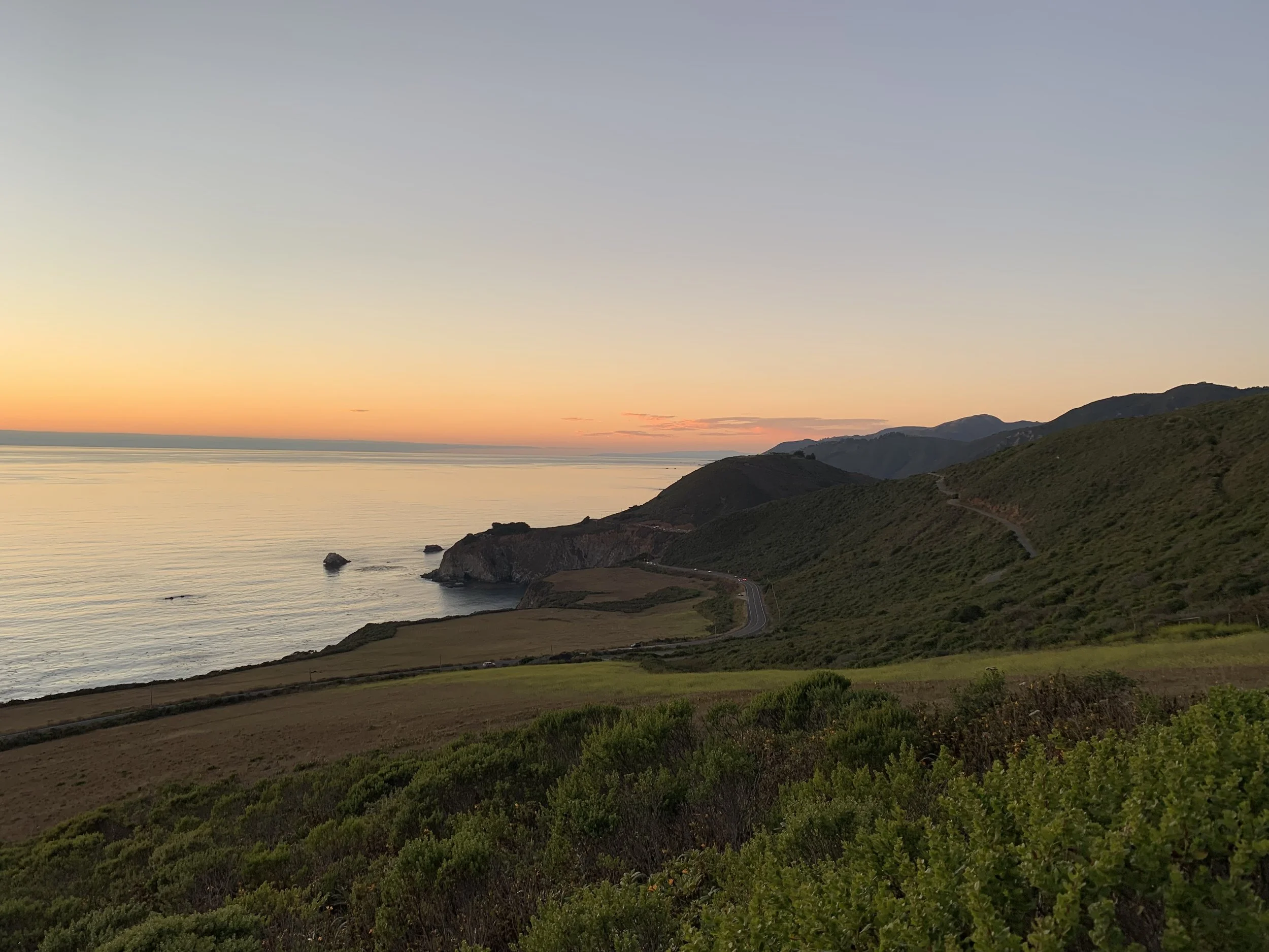 Scenic coastline view of rolling green hills leading to the ocean, with a colorful sunset sky and a few clouds.