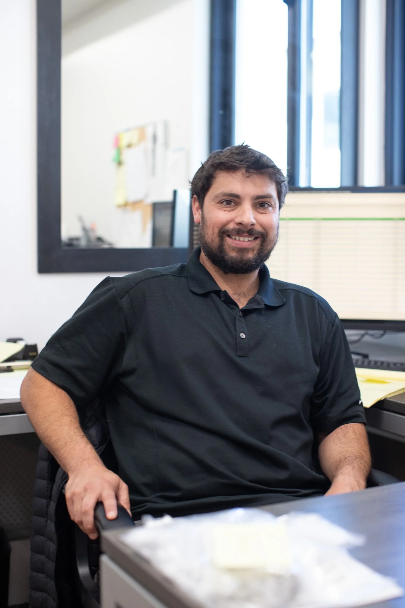 A man with dark hair and a beard smiling, sitting in an office, wearing a black polo shirt. Office background includes a window, computer, and papers on a desk.