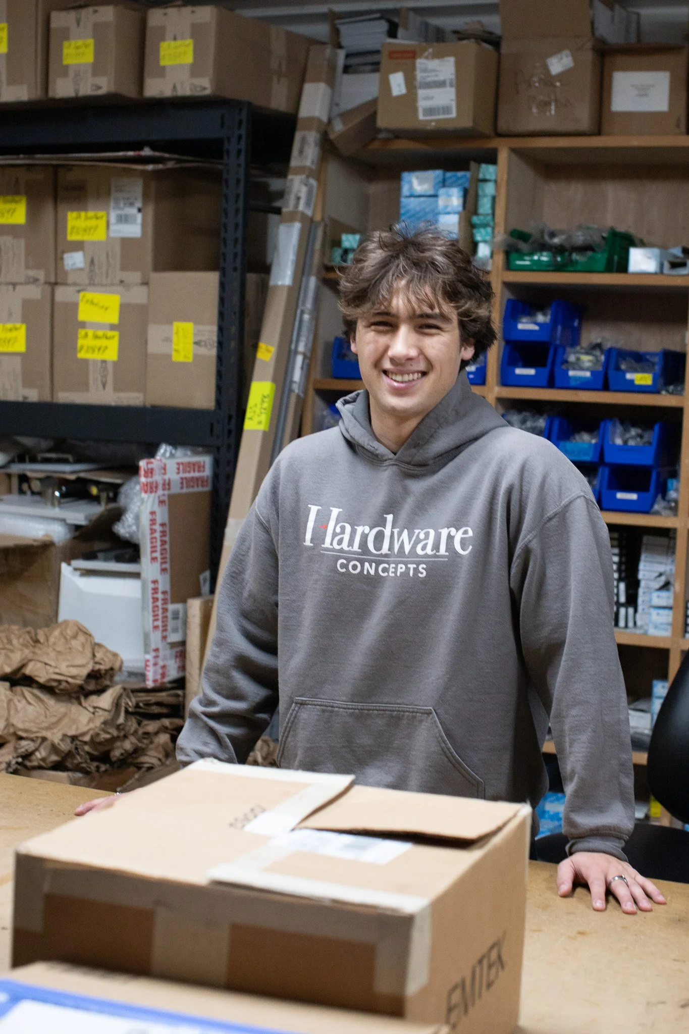 A young man smiling while standing in front of shelves filled with boxes and supplies in a warehouse or storage room.