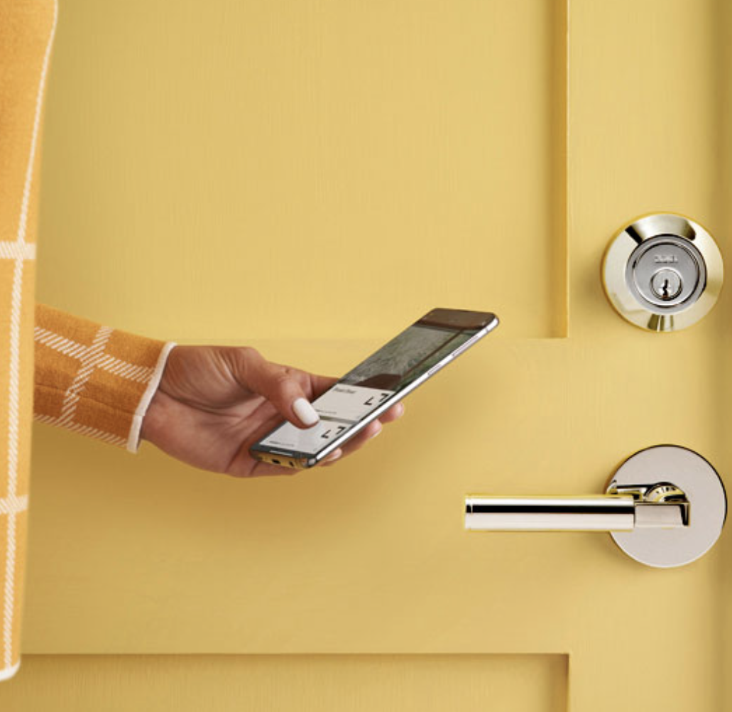 A hand holding a smartphone with a yellow checkered long sleeve, near a yellow door with a door lock and a modern silver door handle.