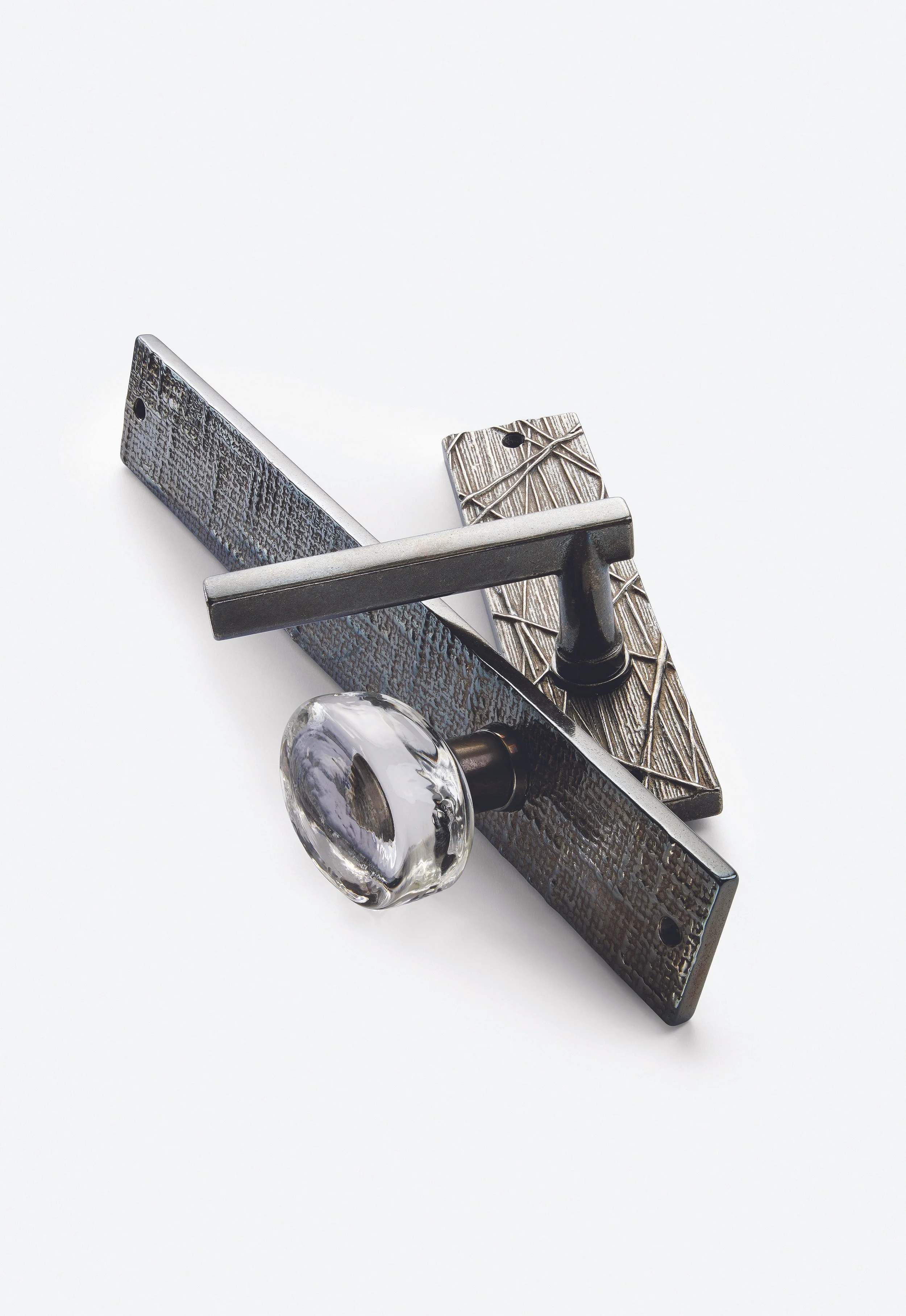 Close-up of a vintage door latch with a textured dark metal backplate, a clear glass handle, and a decorative metal latch with a leaf pattern, all set against a plain white background.