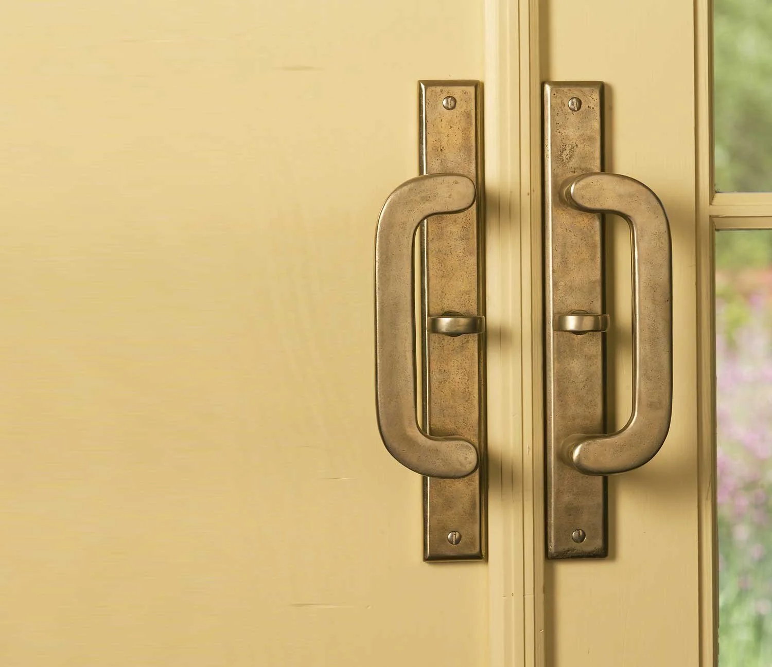 Close-up of yellow door with metal handles and a window showing greenery outside.