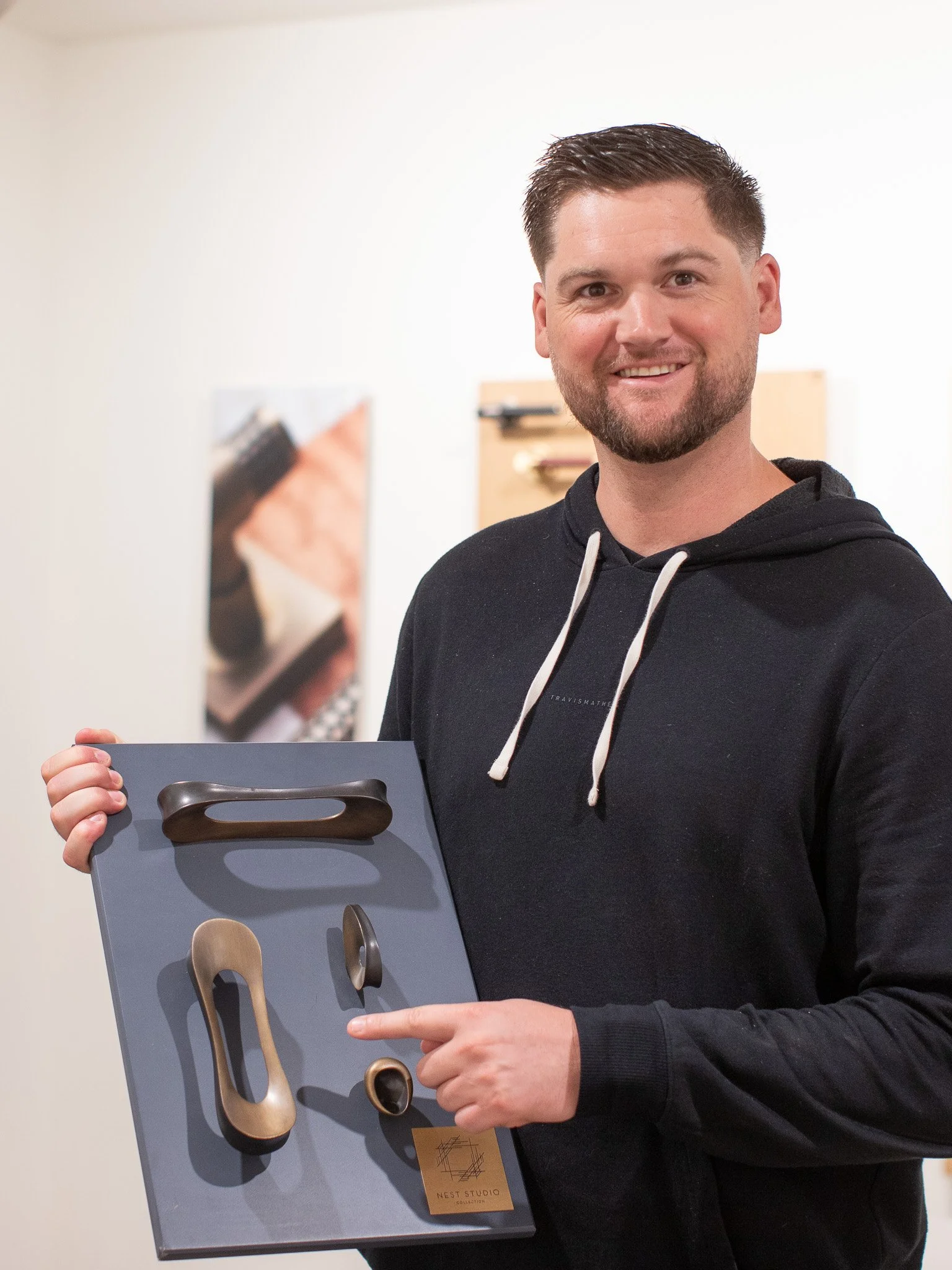 A man with short dark hair and a beard smiling, holding a display board with various modern, abstract door handles. He is wearing a black hoodie with white drawstrings, standing in front of a neutral background.