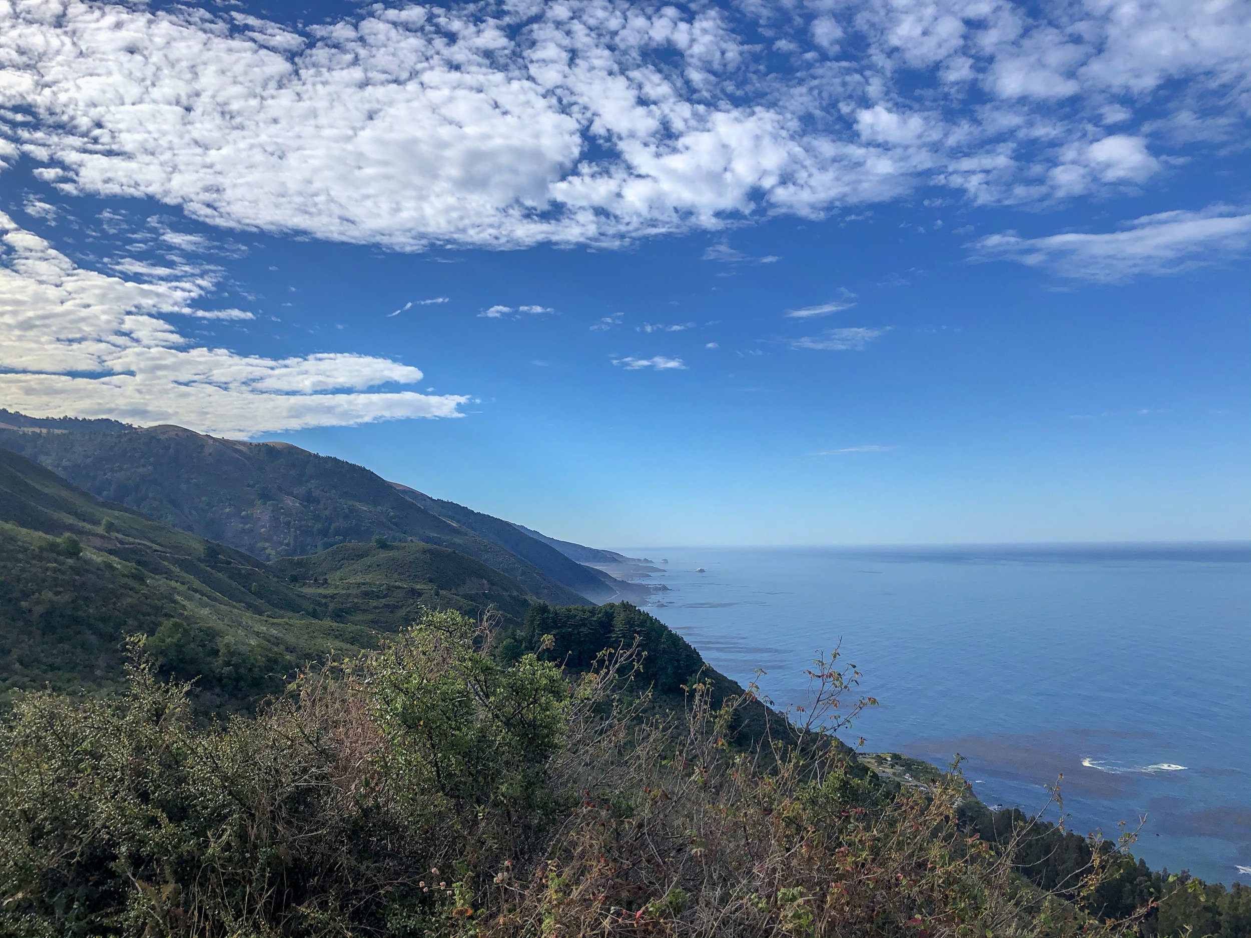 Scenic view of the coastline with mountains and greenery in the foreground, and the ocean extending to the horizon under a partly cloudy sky.