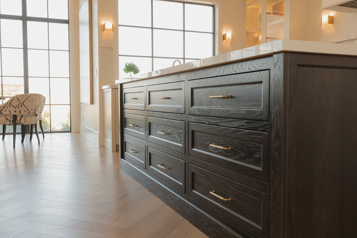 Modern kitchen with a dark wooden island, large windows, light wood flooring, and a dining area with a patterned chair.