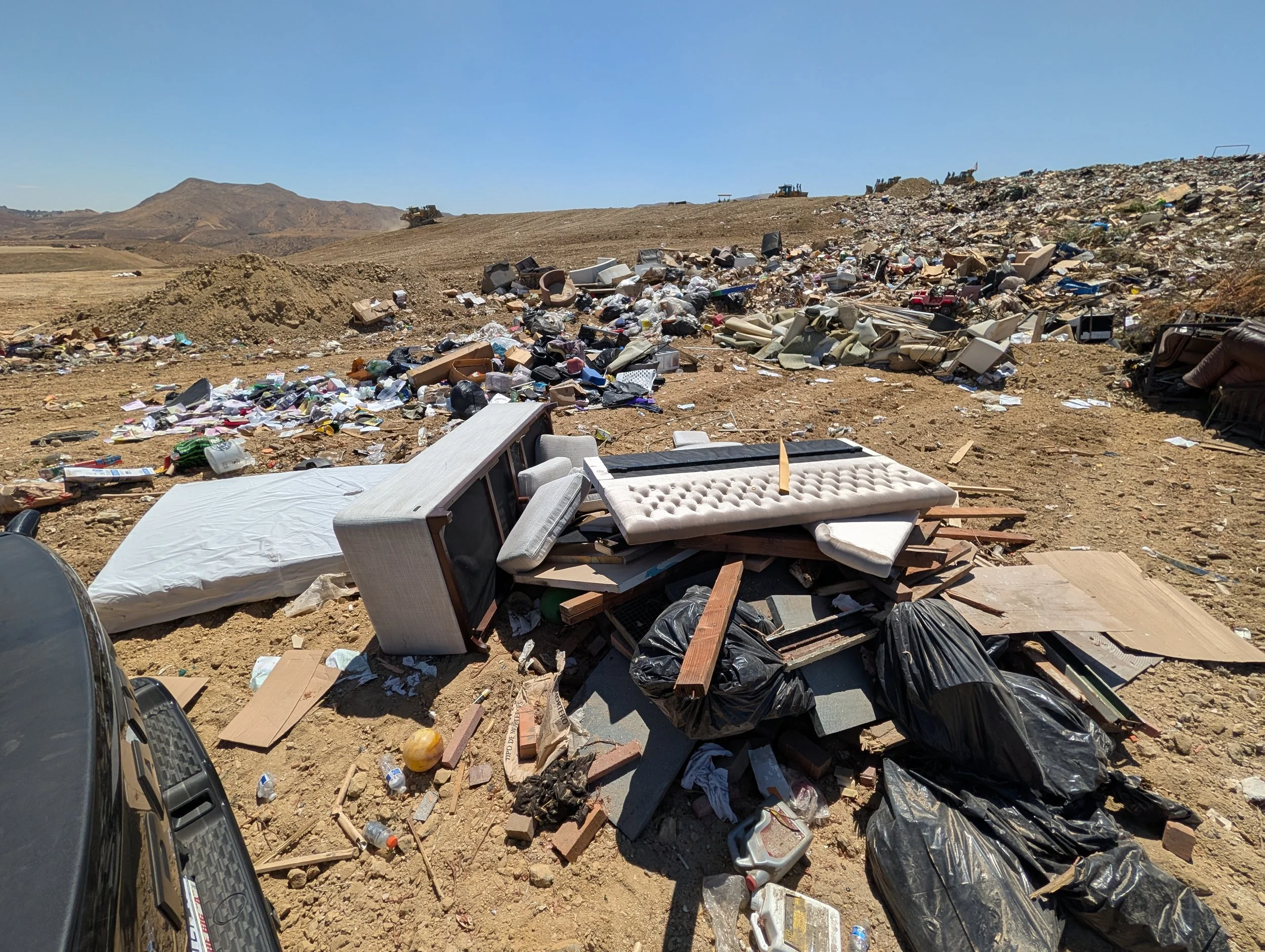 A landfill dumping ground filled with scattered trash including mattresses, plastic bags, boxes, and debris under a clear blue sky with hills in the background.