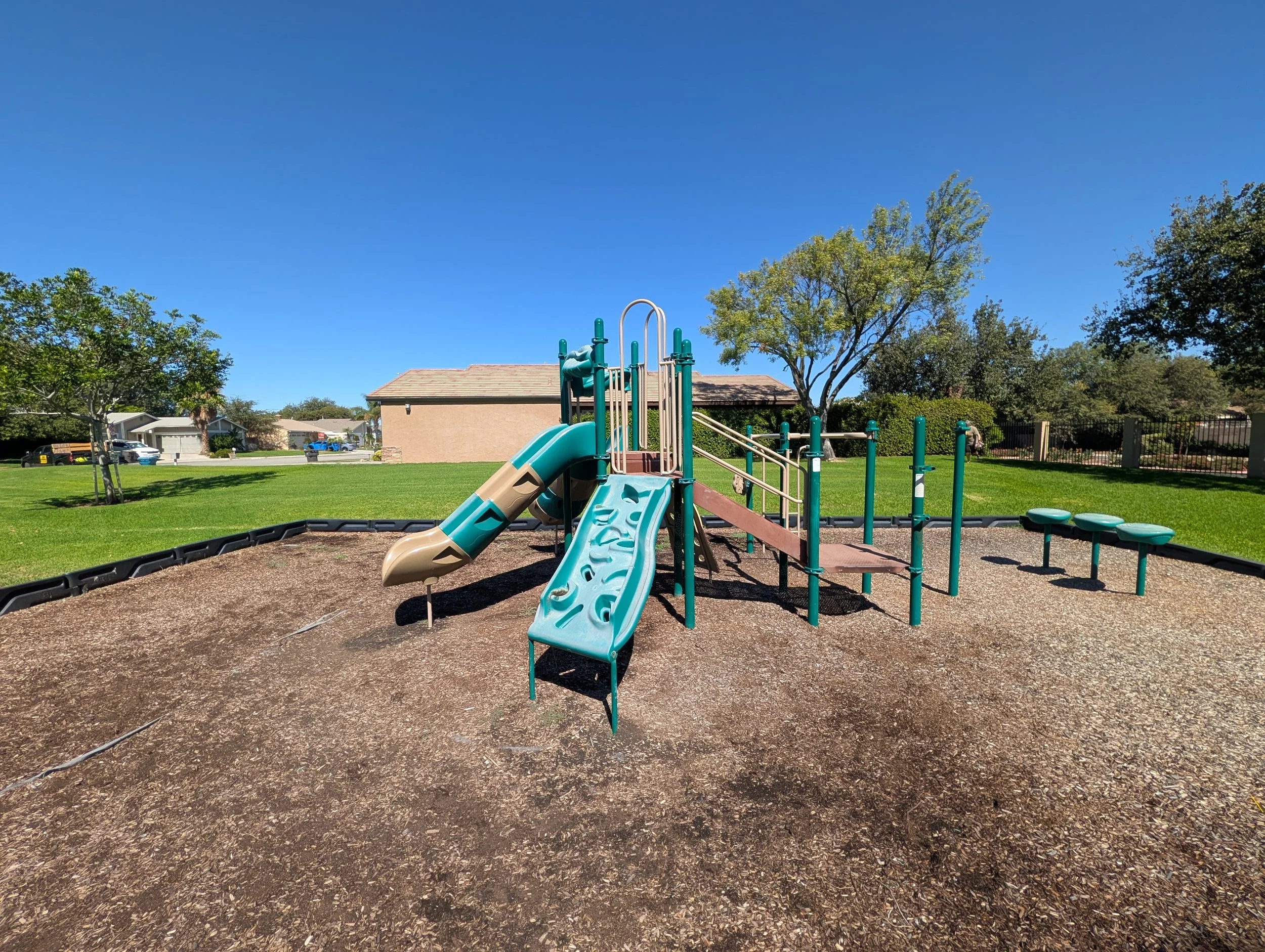 A playground with slides and climbing structures on a grassy field under a clear blue sky, with trees and residential houses in the background.