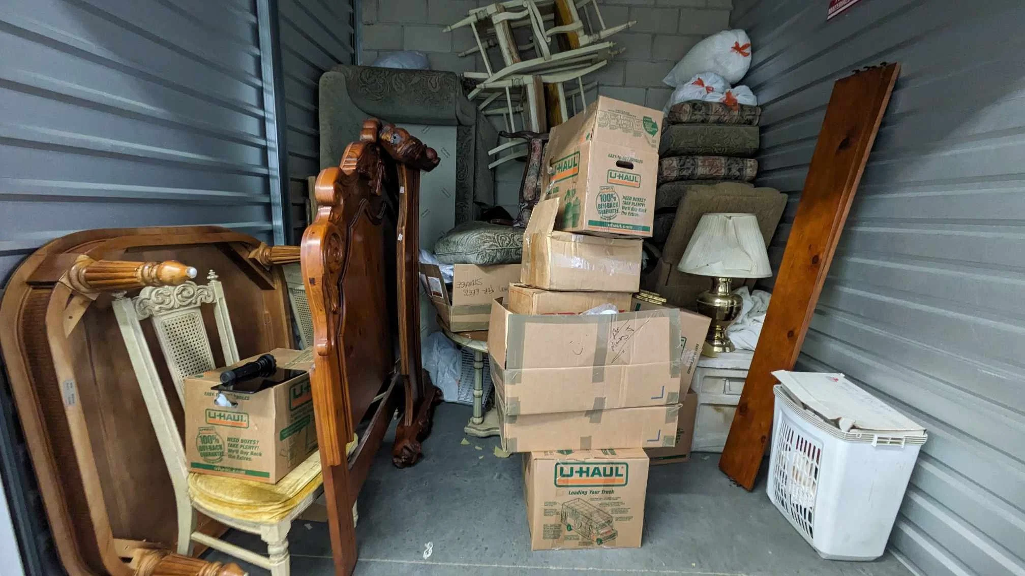 Storage unit filled with furniture, boxes, and household items, including dining chairs, a mirror, and a lamp.