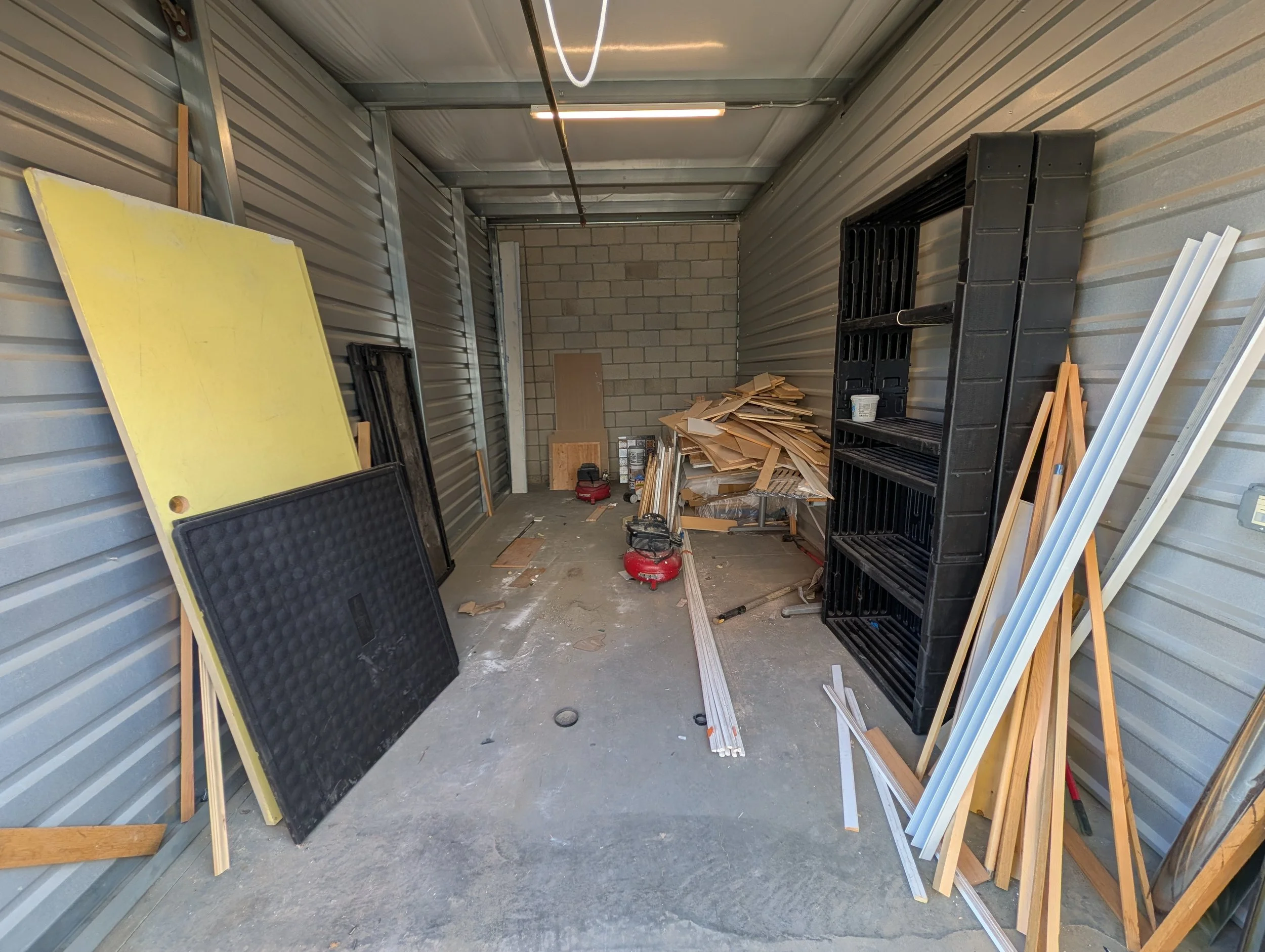 Inside a storage unit with metal walls, construction materials including wood, plastic panels, and tools are scattered around. There are black plastic shelves and a pile of wooden pieces on a table.
