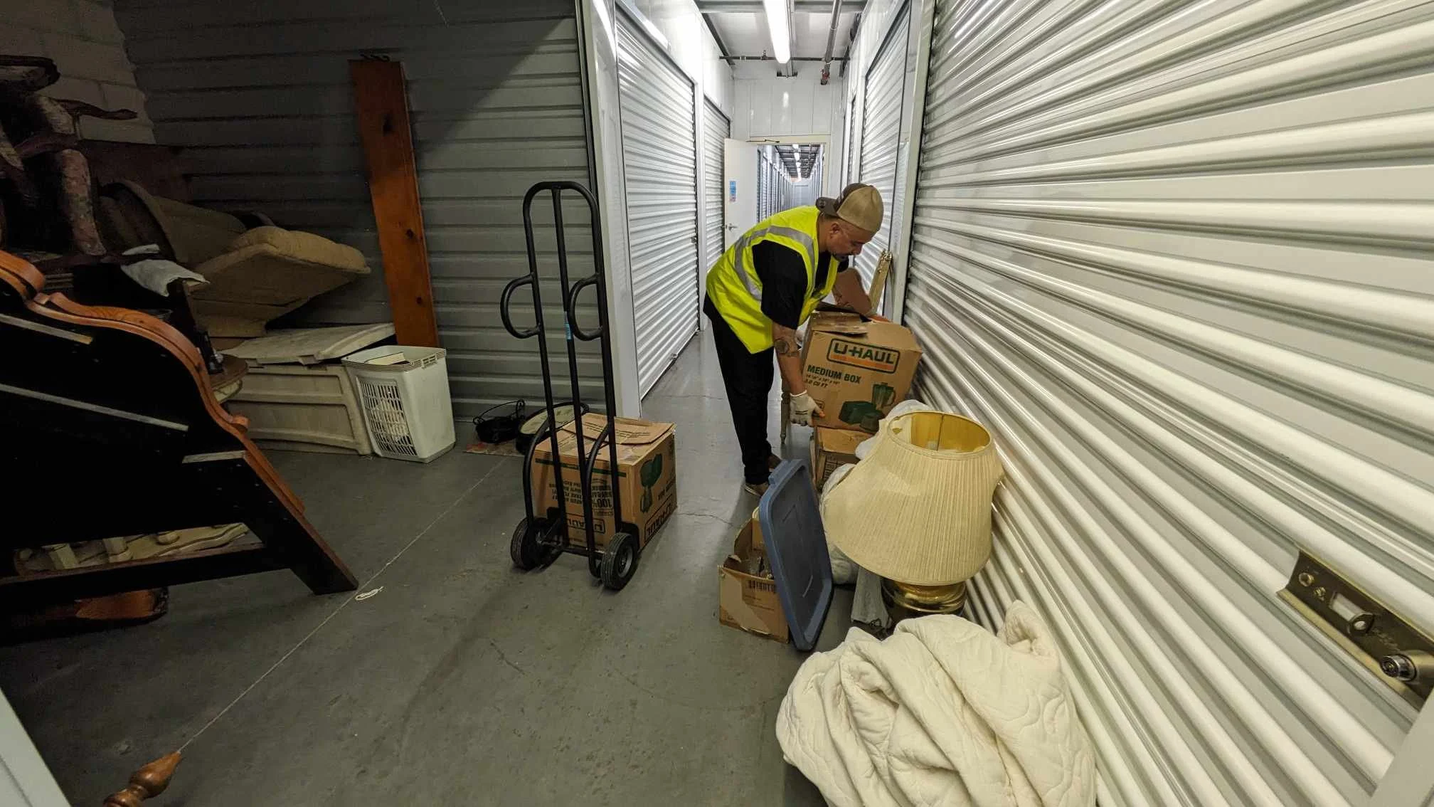 A person with a cap and a high-visibility vest is unpacking at a storage unit with white metal roll-up doors. They are handling a medium-sized box labeled 'U-Haul.' Various boxes and furniture items are around, including a lamp, a lamp shade, an armchair, and a cart.
