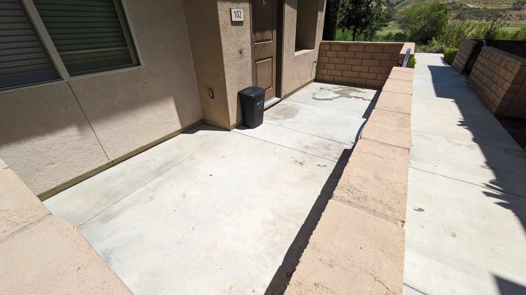 A front porch of a house with a concrete floor, a brown door, and a black trash bin. The house has a beige stucco exterior with a window, and a small brick side wall. There is a railing and scenic green hills in the background.