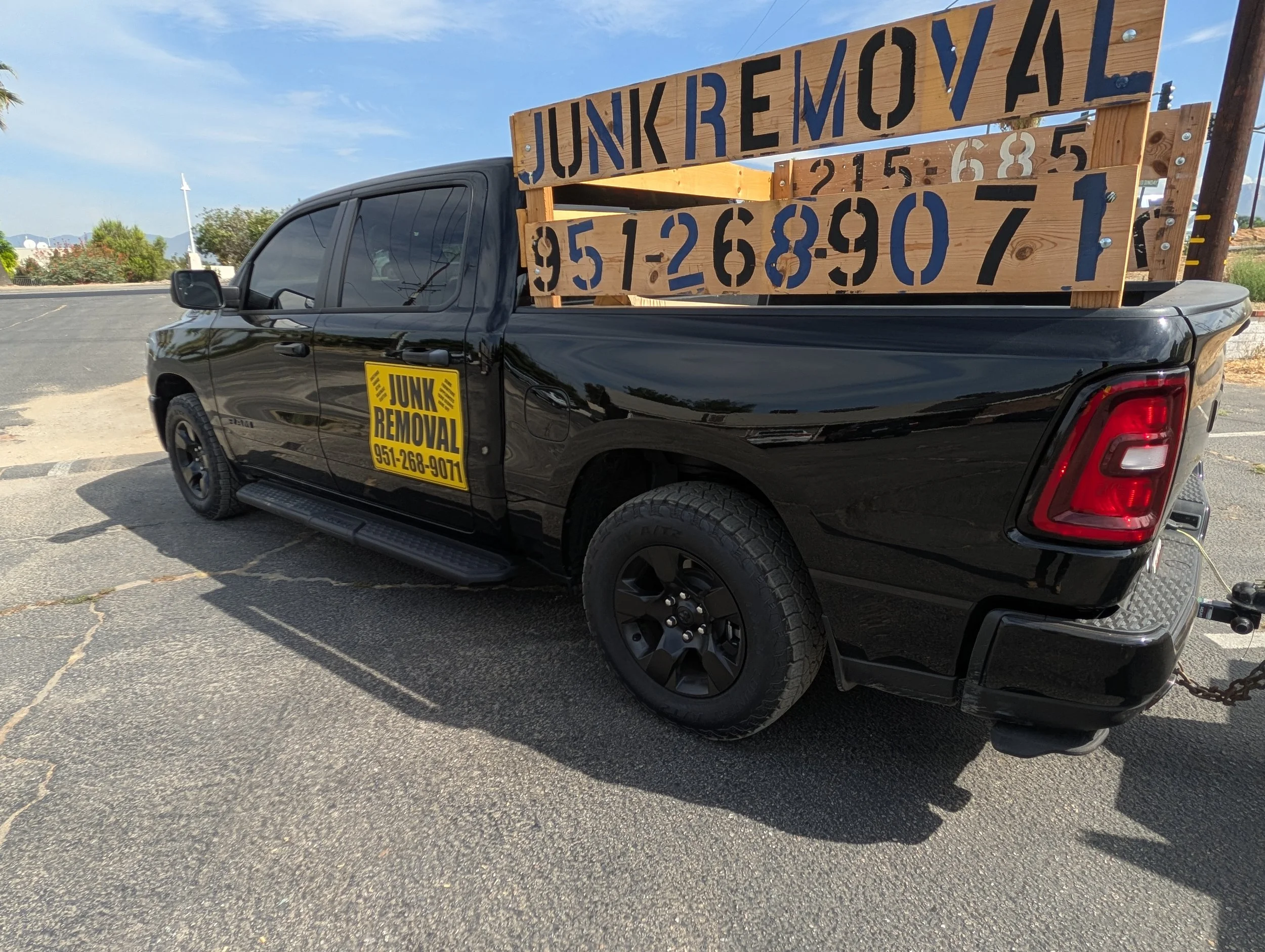A black pickup truck with a yellow sign on the door that reads 'Junk Removal' and a phone number, and large wooden signs on the bed of the truck with text about junk removal services and phone numbers, parked on asphalt pavement with a partly cloudy sky and some trees and buildings in the background.