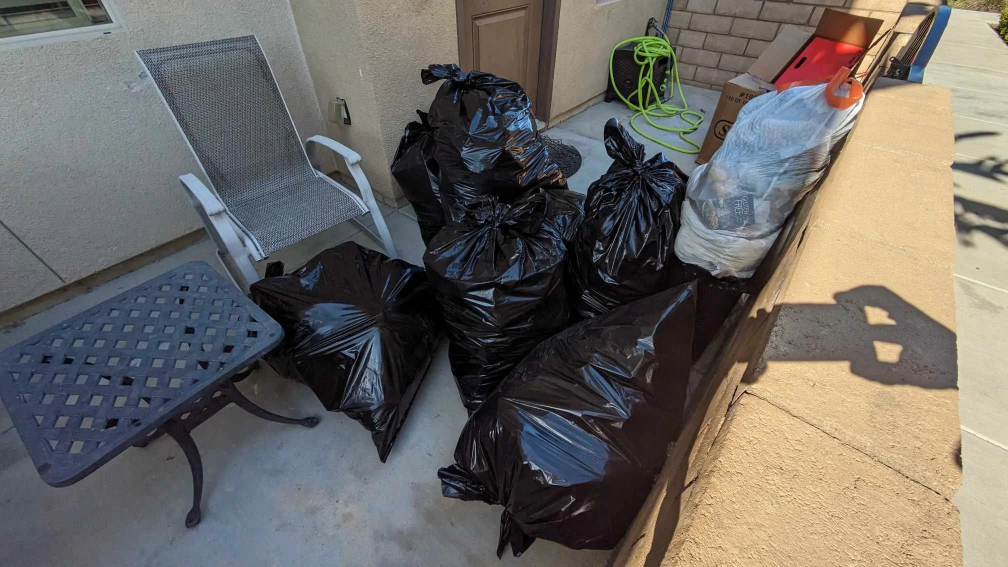 Several black trash bags on a porch next to a metal outdoor table and chair, with a green garden hose coiled nearby.