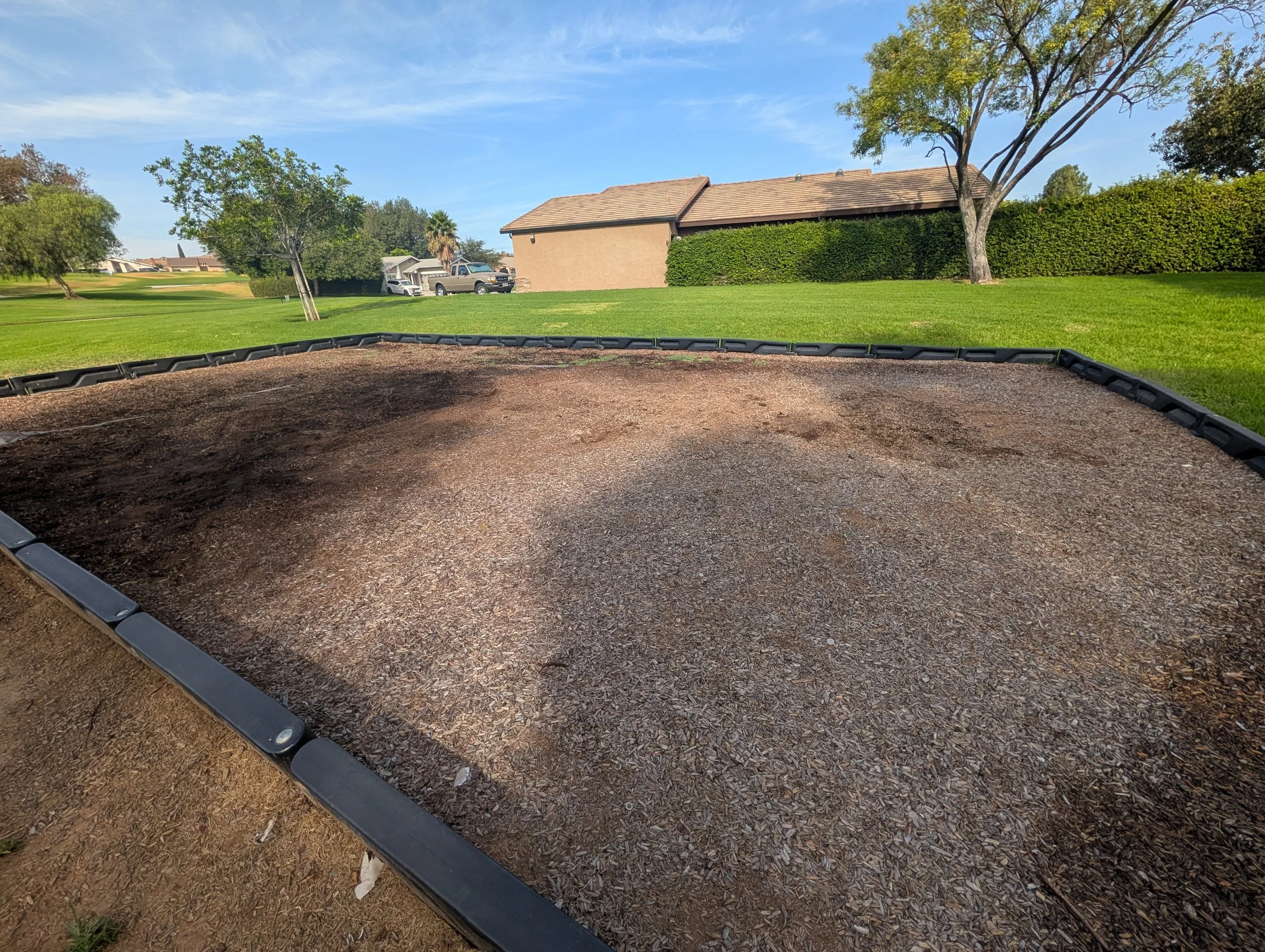 Empty animal playground sandbox with a black border on green grass, with trees, houses, and cars in background under a blue sky with some clouds.