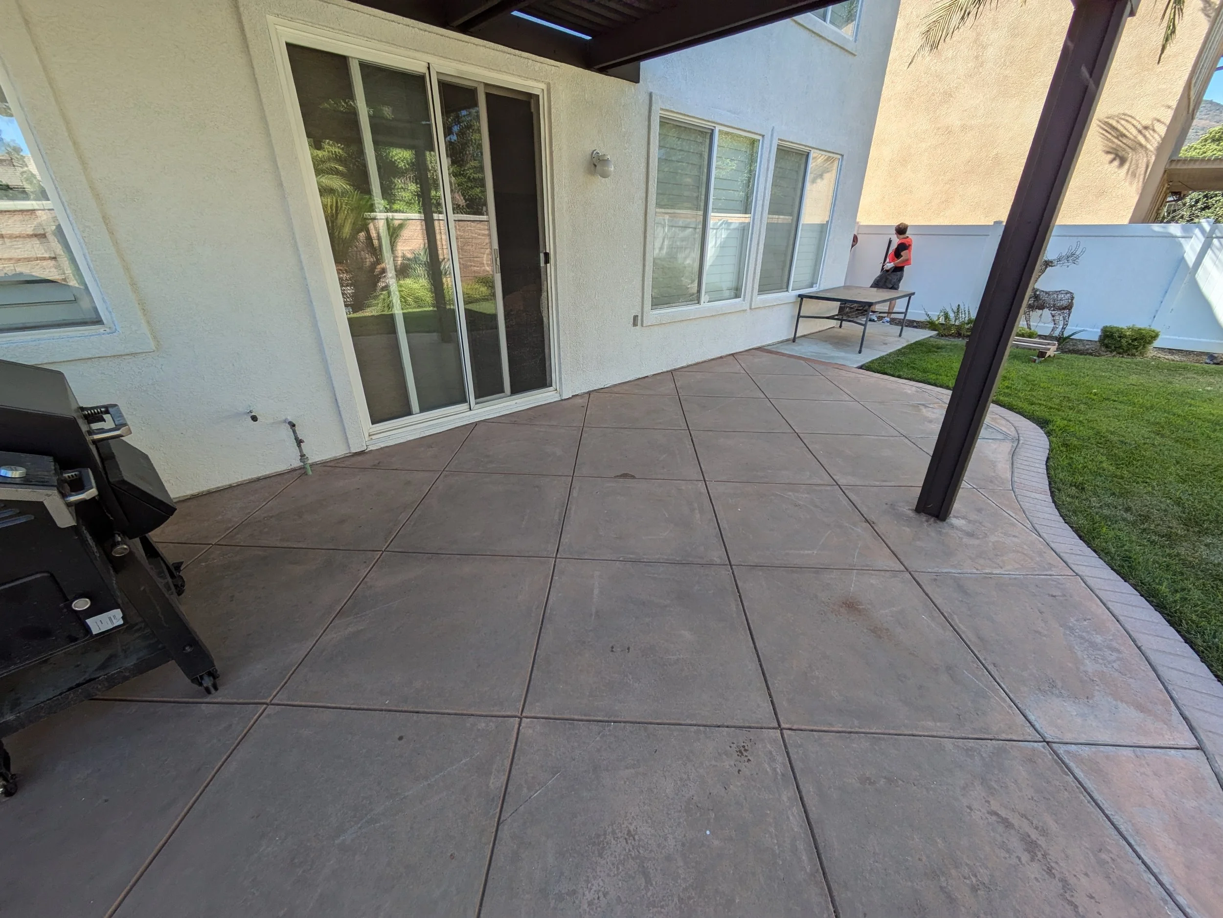 A backyard patio with a sliding glass door, a table, a grill, and a person walking, surrounded by a lawn and garden with a privacy fence.