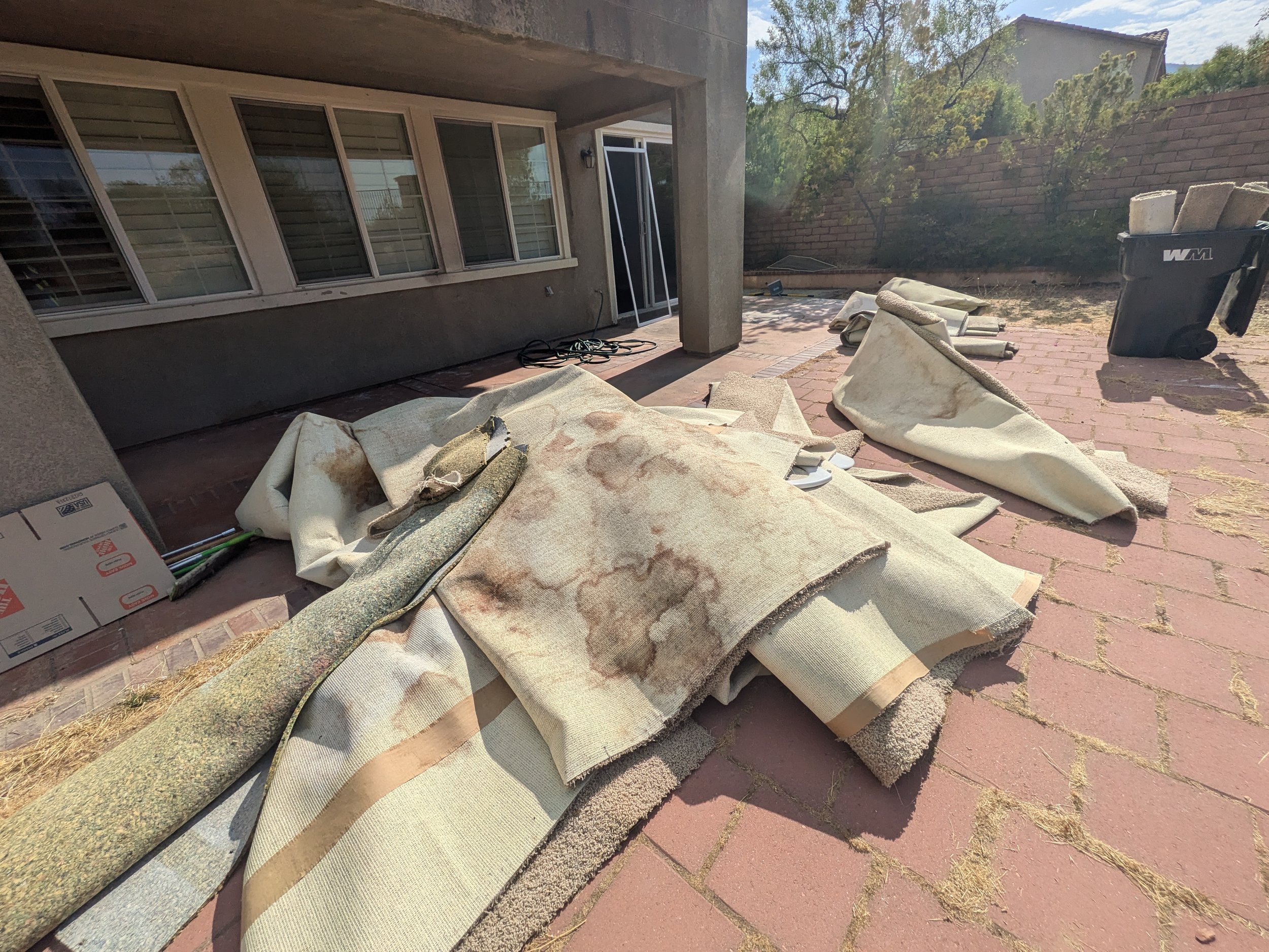 Backyard patio under renovation with rolled carpets, a garbage bin, and construction materials scattered on a brick surface, with a house wall and a brick wall with trees in the background.