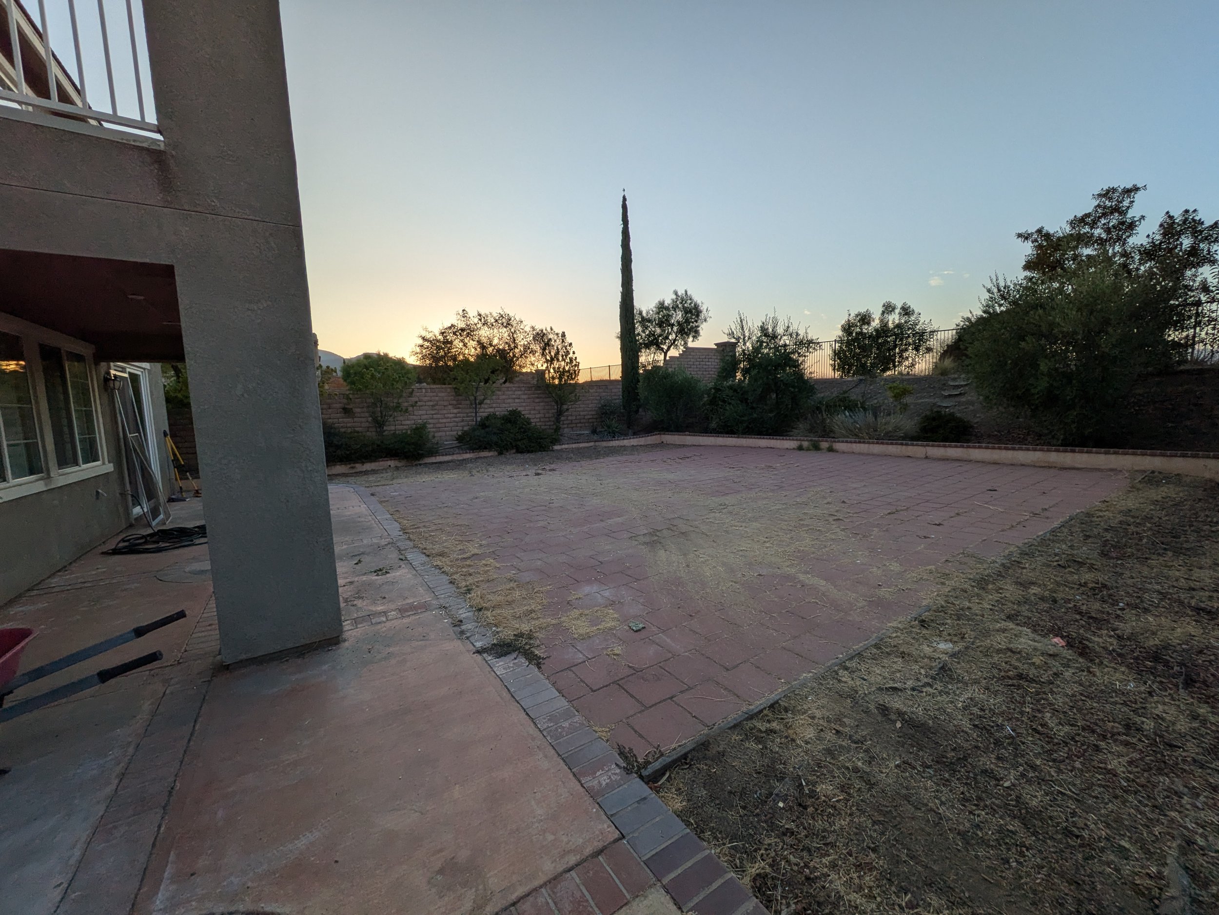 Backyard patio area with brick pavers, a hot tub, and landscaping including trees and bushes. The sky is clear with a slight sunset glow.