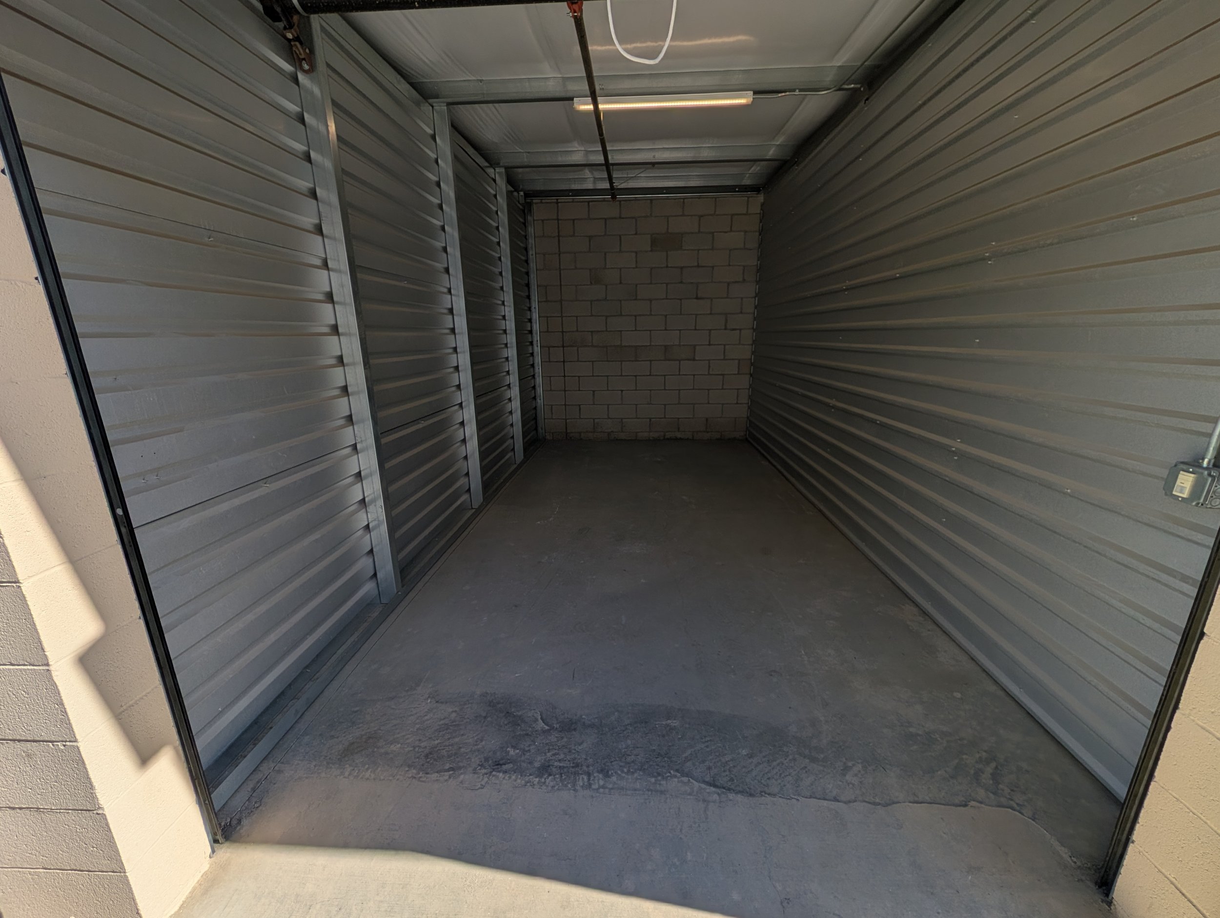Empty storage unit with metal walls, concrete floor, a brick wall at the back, and a ceiling light fixture.
