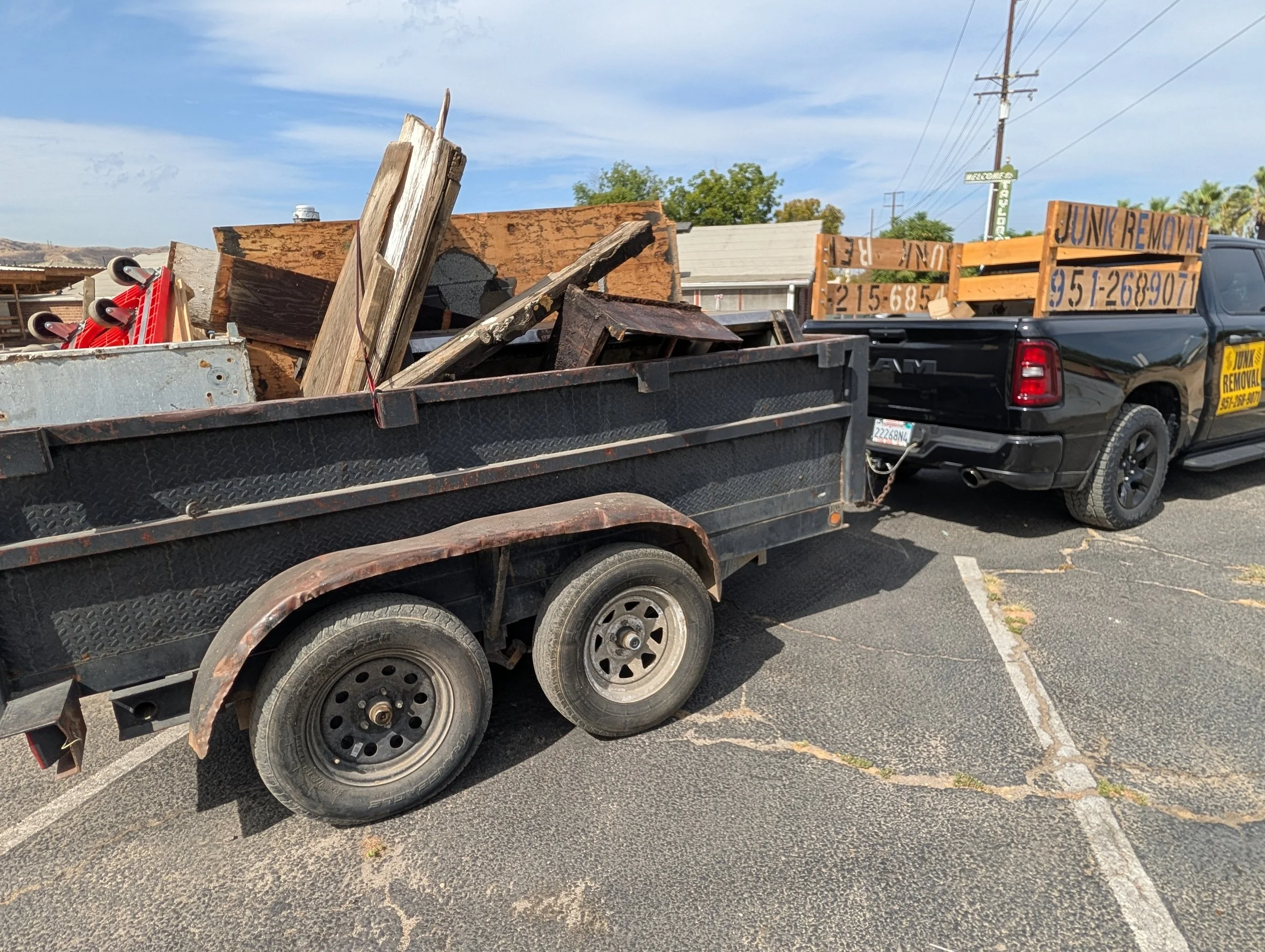 A black pickup truck with a yellow and black sign on the side that reads 'Junk Removal' and a phone number. The truck has a wooden sign with the same text and phone number. The truck bed is filled with various debris, including wood, metal, and other materials. The truck is parked in a lot with a house and trees in the background, and utility poles and wires above.