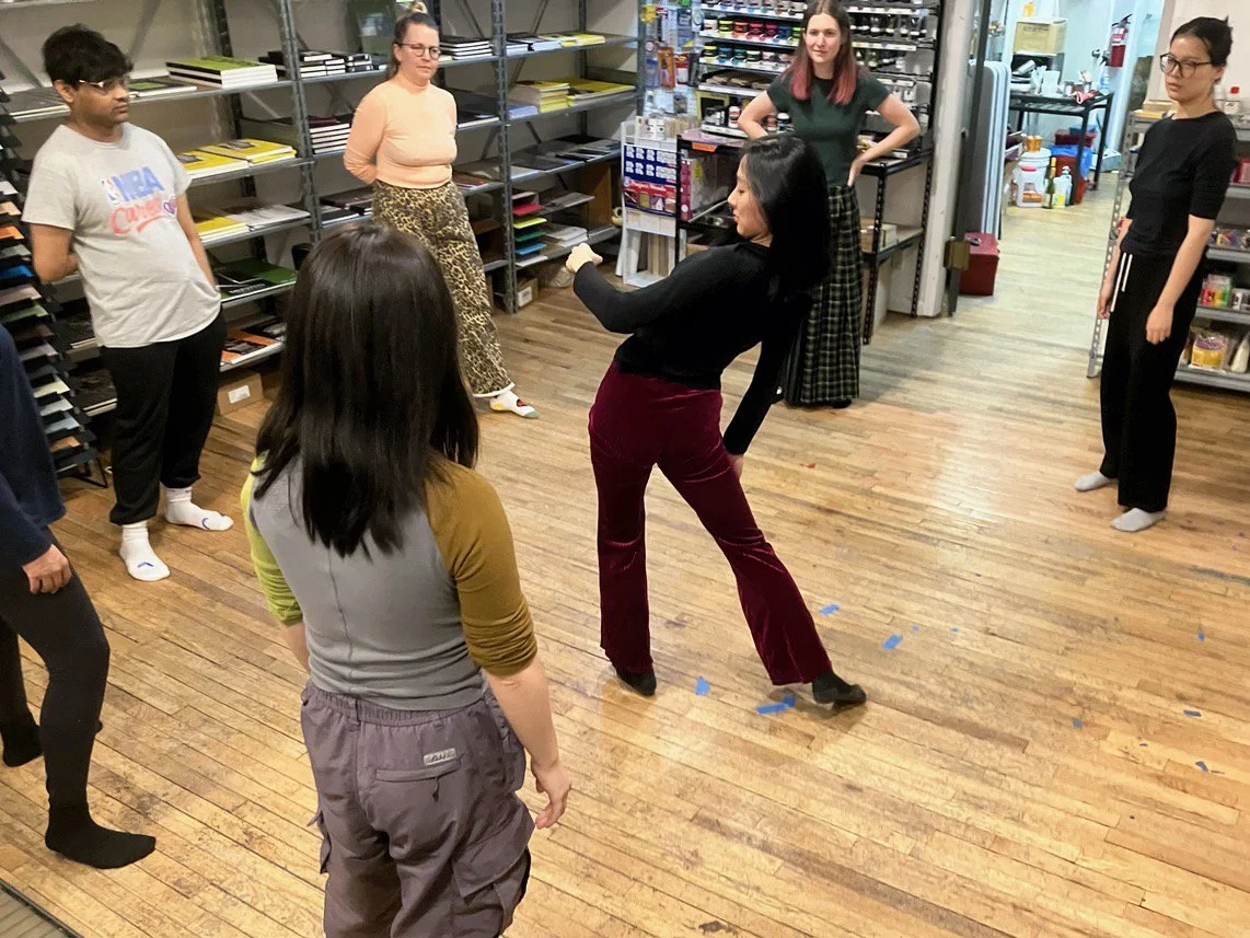 A group of women and a girl in a workshop or classroom setting with shelves of papers, arts, and crafts supplies. One woman is demonstrating a pose or dance move as others watch.