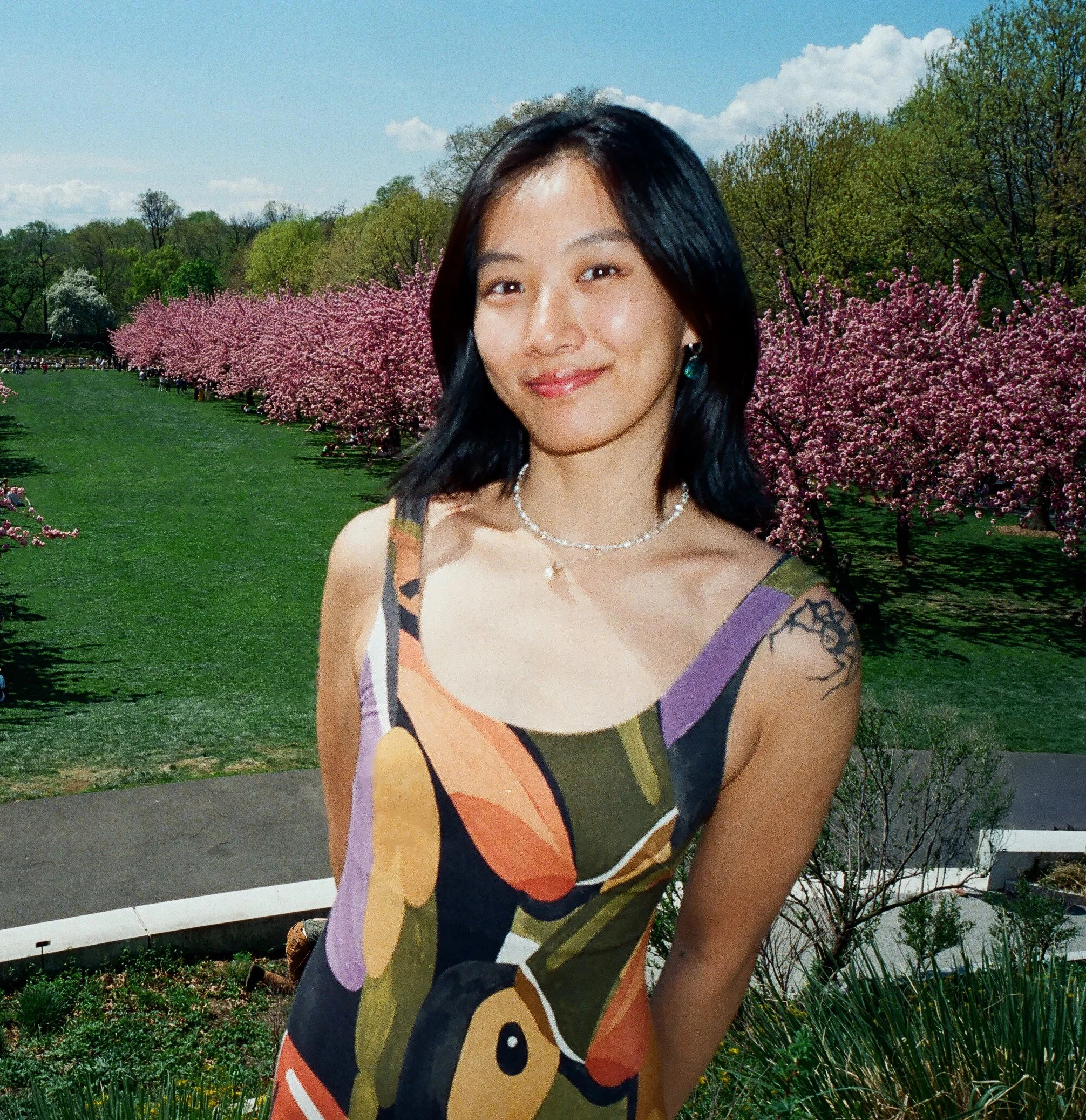 Close-up of a smiling woman with black hair outdoors in front of flowering trees and a cloudy sky.