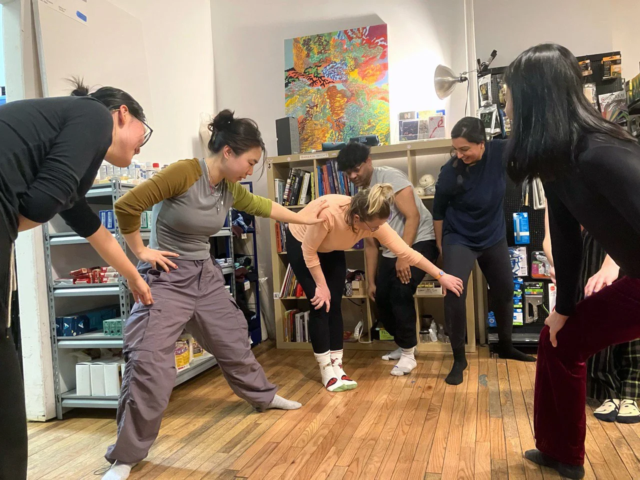 Group of seven women participating in a stretching or yoga session in a room with bookshelves and a colorful painting on the wall.