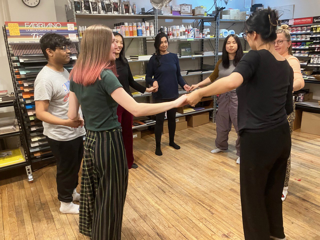 A group of women and one young man are standing in a circle, holding hands, in a store or office with shelves stocked with office supplies and art materials.