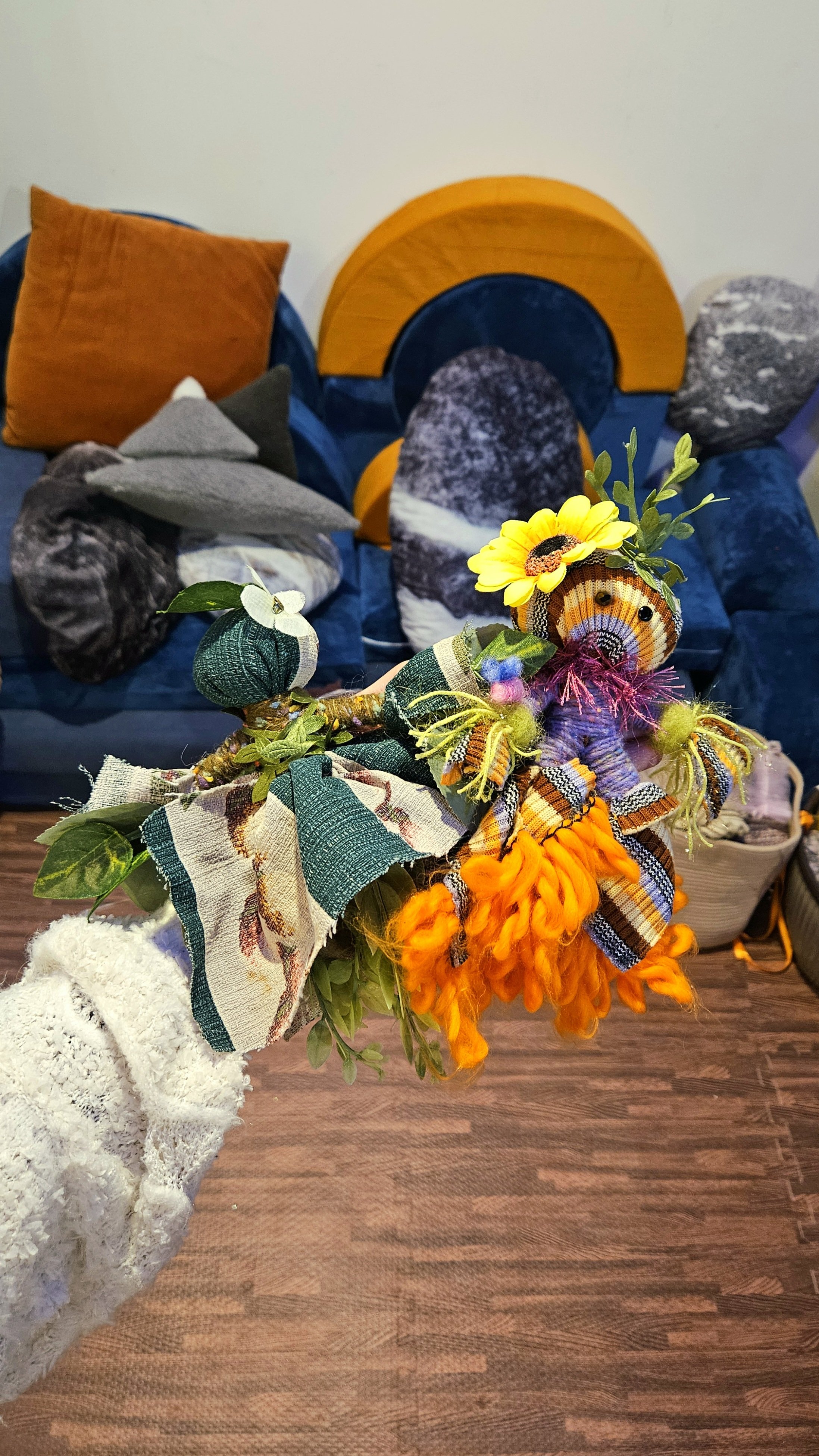 A person wearing a white fluffy sweater holds a bouquet of fabric and artificial flowers, including sunflowers and autumnal leaves, in front of a blue velvet couch decorated with colorful pillows and a semi-circular wall hanging.