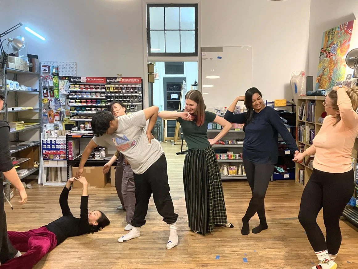 Group of six women engaging in playful activity or game in a bookstore or library, with shelves of books, art supplies, and a whiteboard visible in the background.