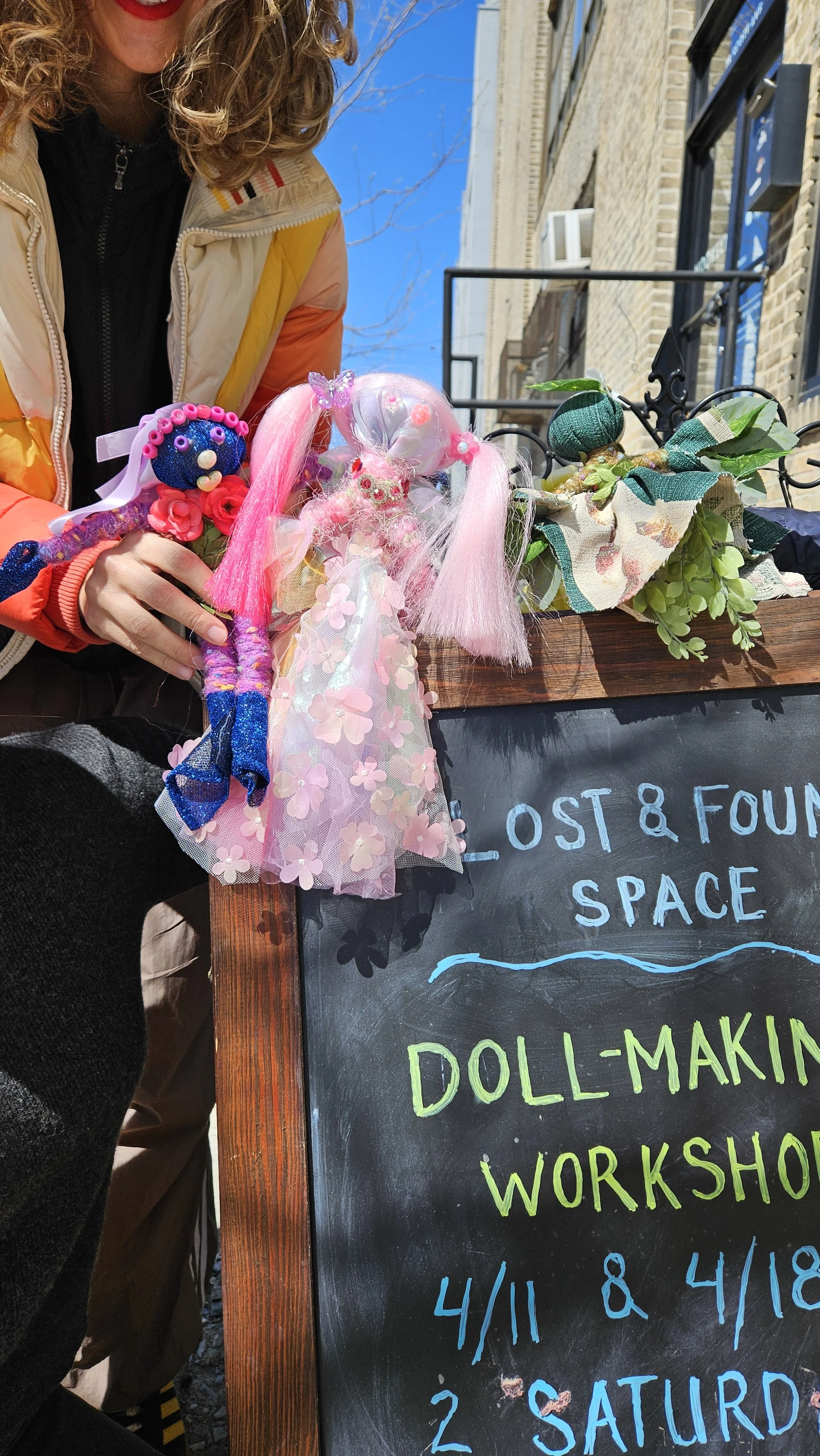 A woman displaying colorful handmade dolls and crafts outside a storefront with a blackboard sign advertising a doll-making workshop on April 11 and 18, with detailed brick building and blue sky in the background.