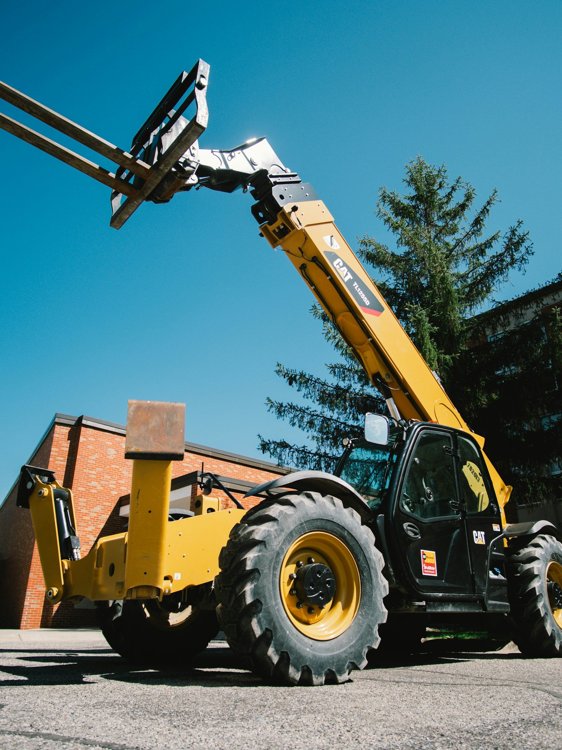 A yellow Caterpillar telescopic forklift with black cab, large tires, and extended arm, lifting a construction or sign post, set against clear blue sky with a tree and brick building in the background.