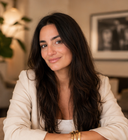 A woman with long dark hair smiling, sitting indoors with a blurred background of framed art and a potted plant.