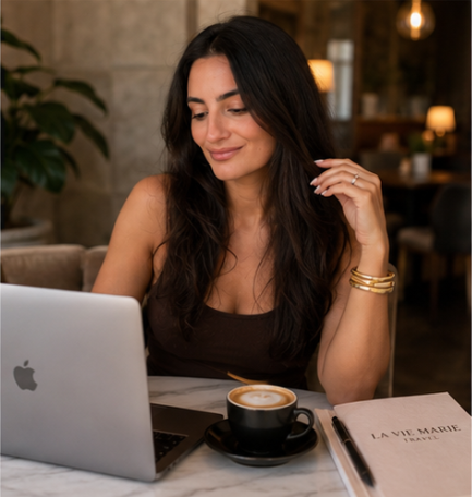 A woman with long dark hair sitting at a table in a cozy cafe, working on her laptop with a cup of coffee and a magazine nearby.