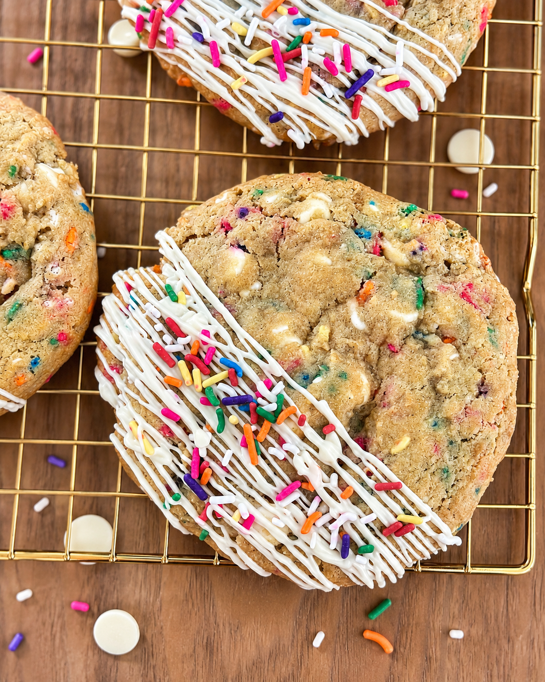 Close-up of two large rainbow cookie bars decorated with white icing and colorful sprinkles resting on a gold cooling rack on a wooden surface with scattered sprinkles and white chocolate chips.