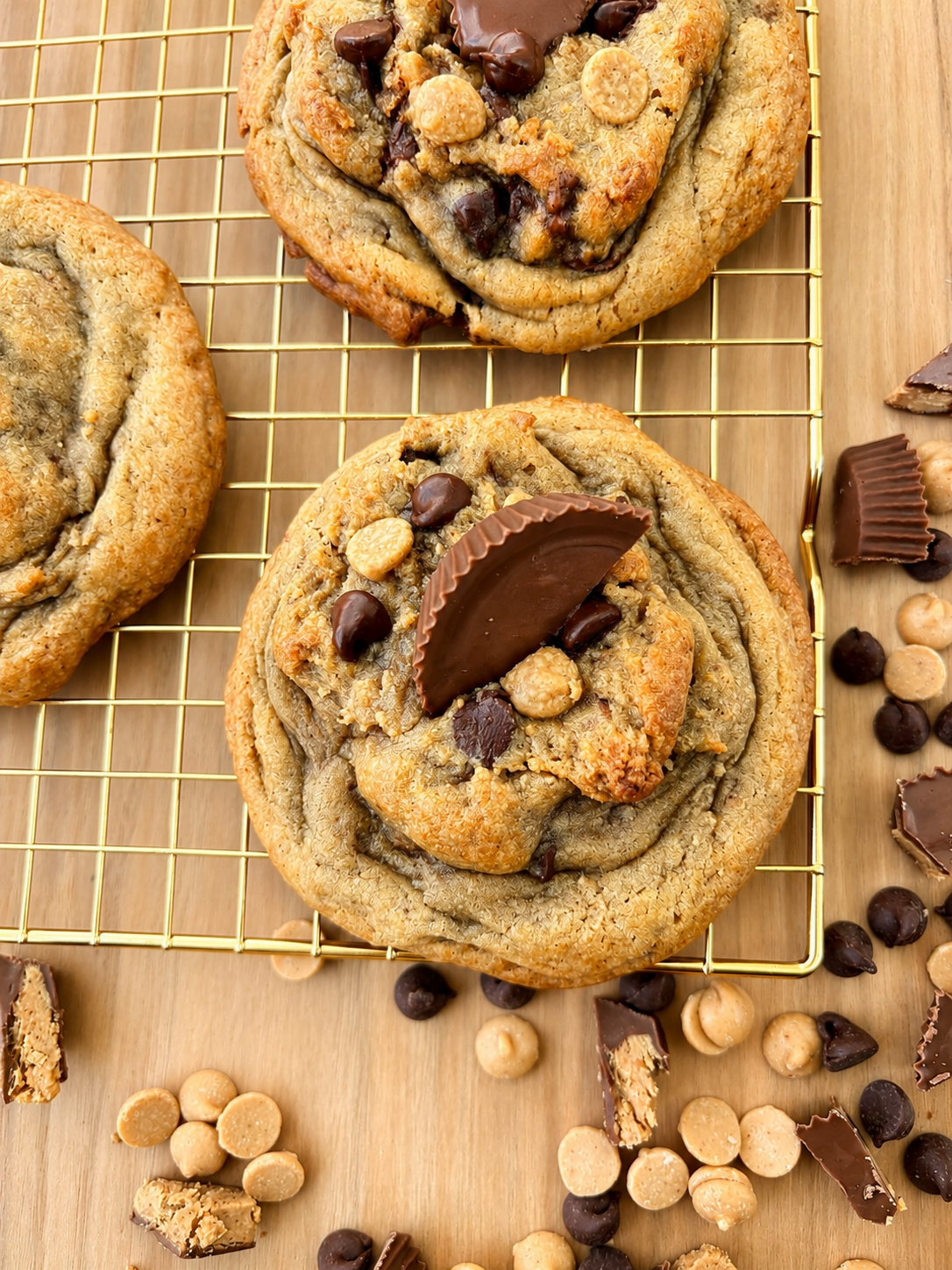 Two chocolate chip cookies topped with a chocolate-chunk candy and scattered chocolate chips and white chocolate chips on a wooden surface.