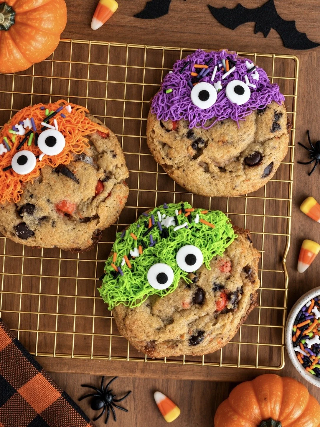 Three Halloween-themed cookies decorated with green, purple, and orange icing, made to look like monster faces with googly eyes and sprinkles, on a wire cooling rack surrounded by small pumpkins, candy corn, and fake black spiders.