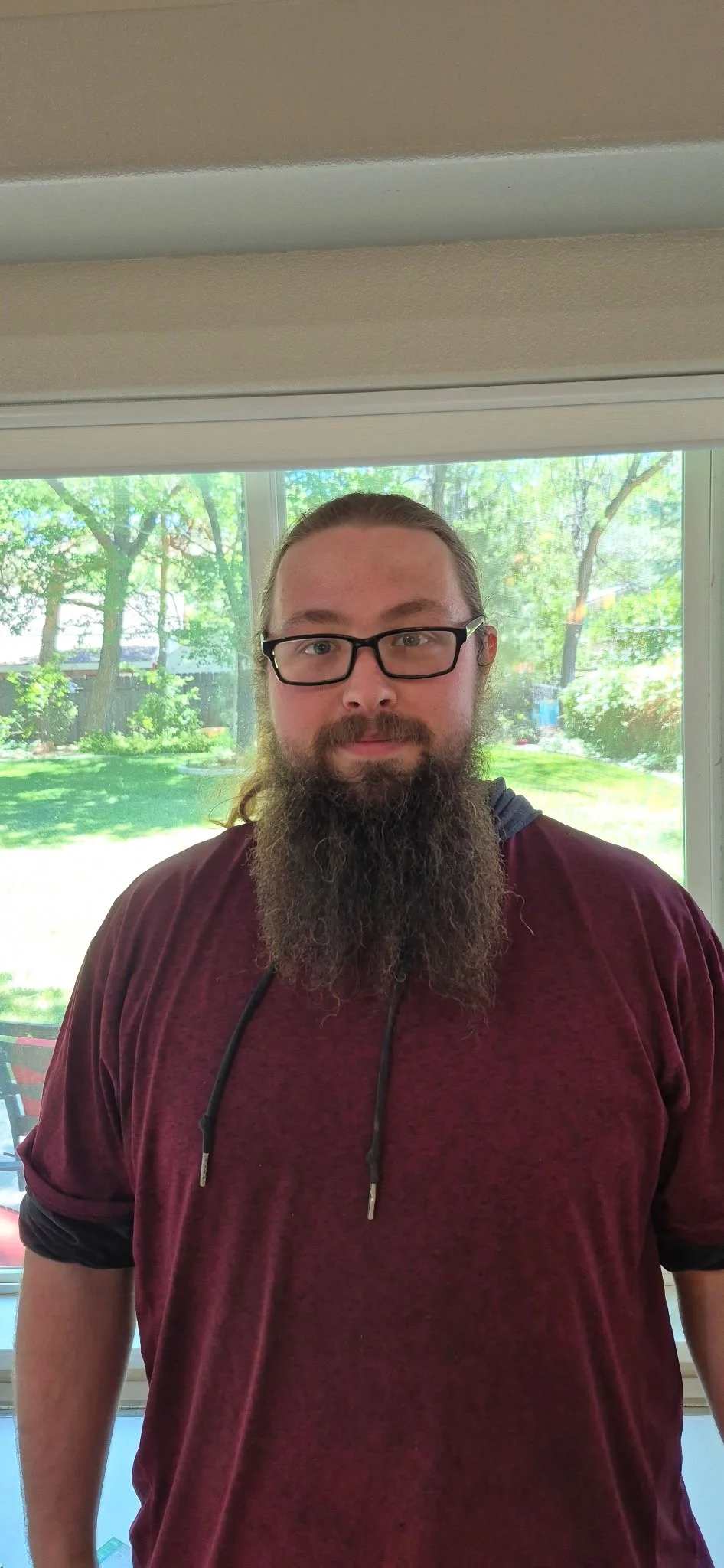 Man with long hair, beard, glasses, wearing a maroon shirt, standing indoors in front of a window with a view of trees and a grassy backyard.