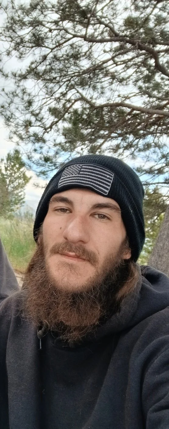 A man outdoors wearing a black beanie with an American flag patch, surrounded by trees and cloudy sky.