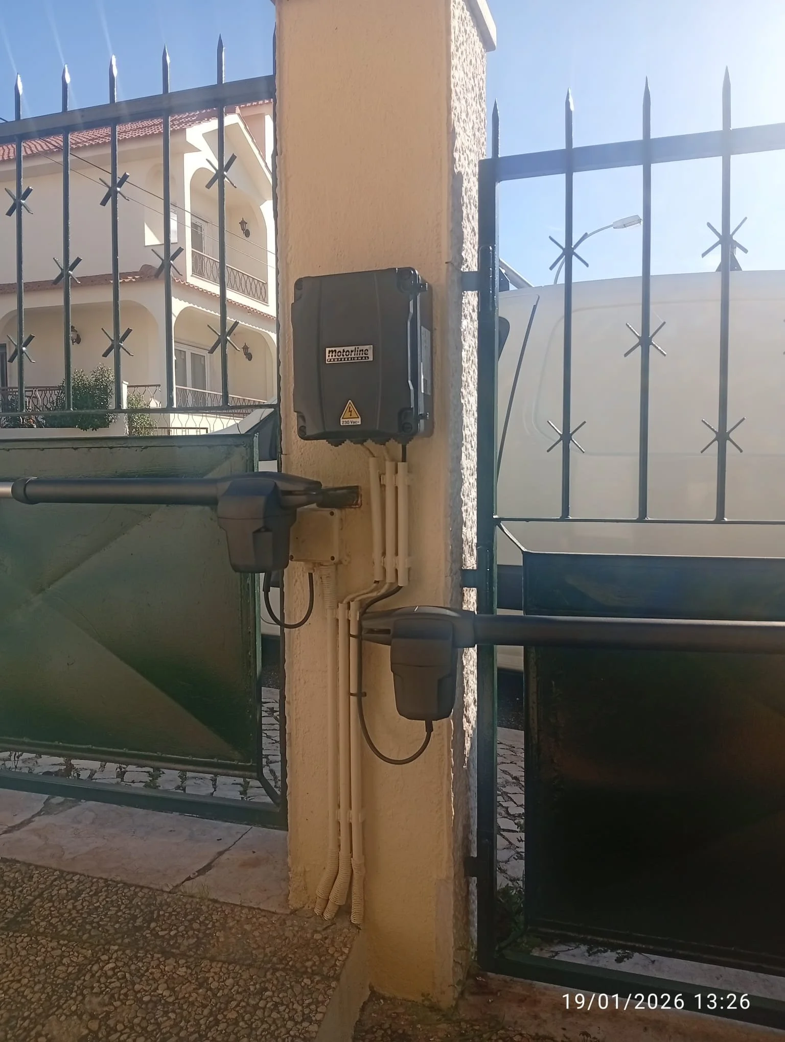 Imagem mostra uma cerca de ferro azul, uma caixa elétrica no muro de uma residência, uma rua de paralelepípedos e uma casa branca com varanda, com o céu ao fundo.