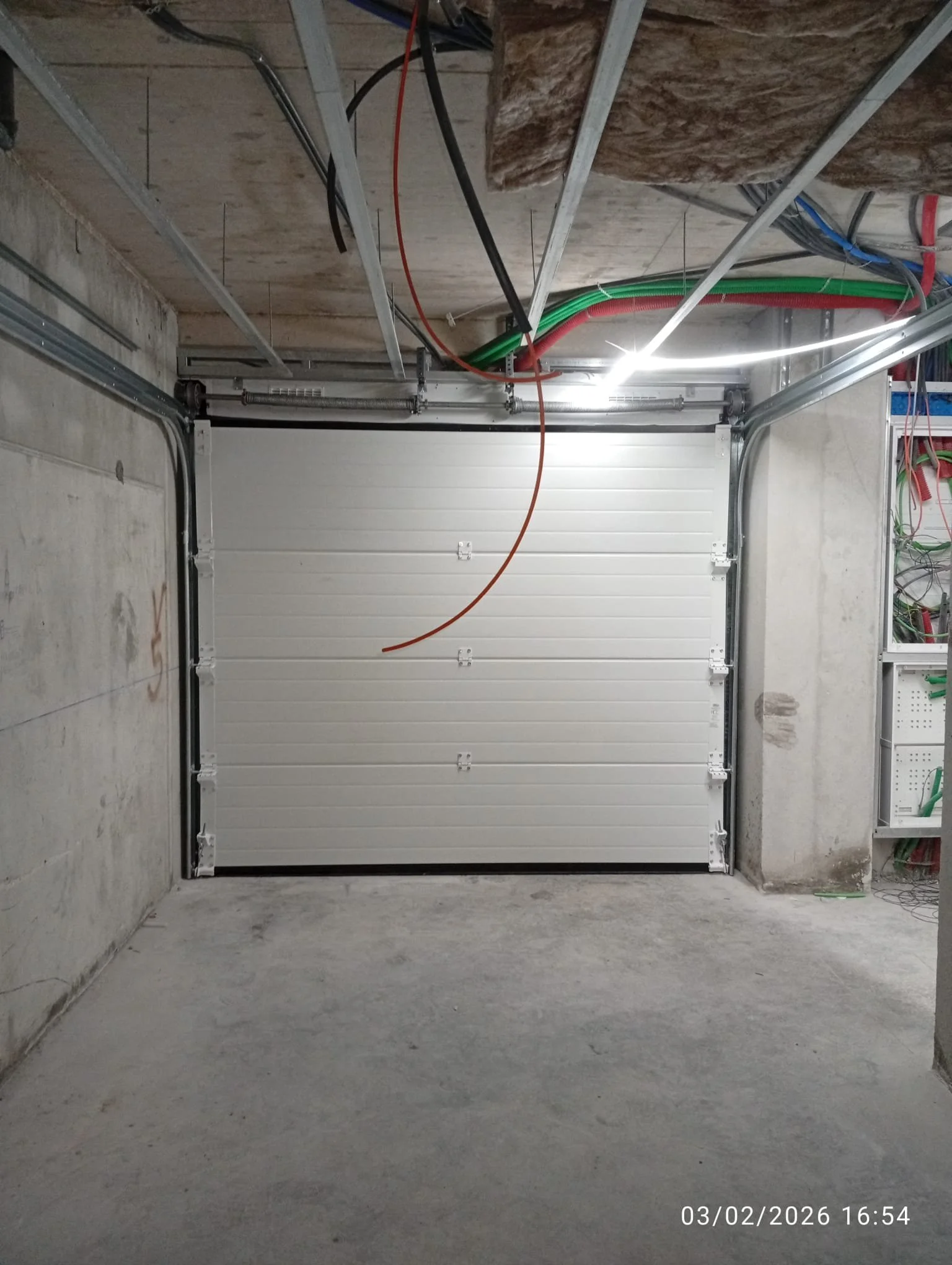 Porta de garagem de aço branca em um ambiente de construção, paredes de cimento e teto com isolamento e cabos de energia visíveis