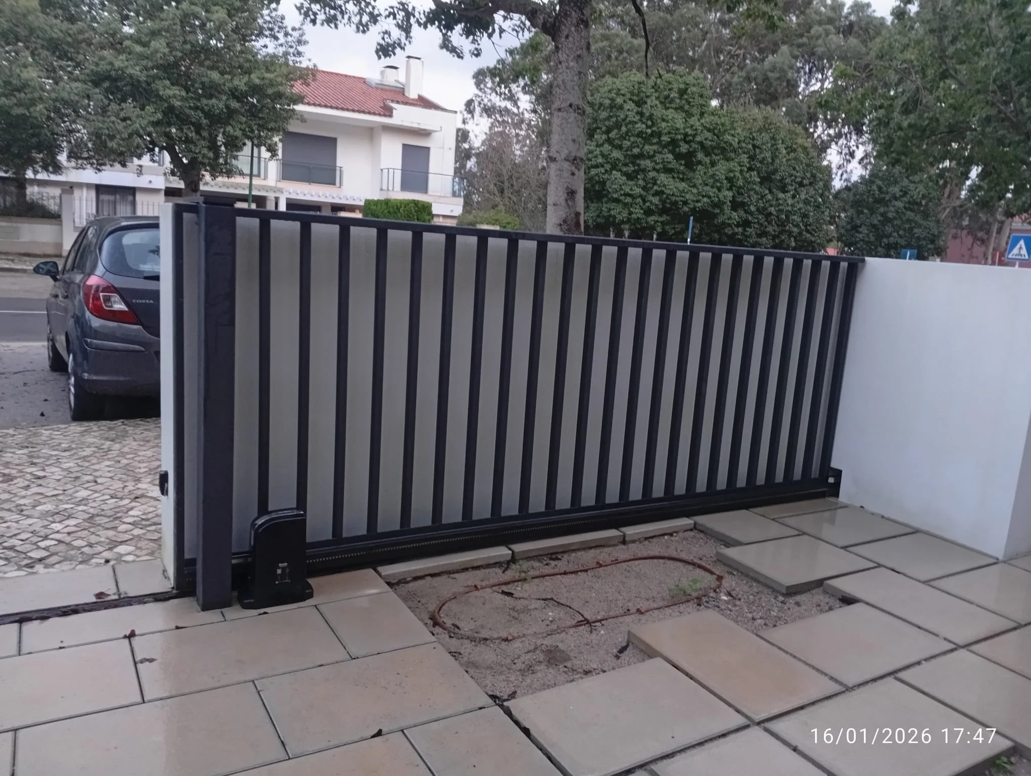 Portão de ferro preto e cinza parcialmente aberto na frente de um jardim com piso de lajotas. Ao fundo, uma casa branca de dois andares com varanda e telhado vermelho, árvores e céu nublado. Vida útil das informações do passado, com data e horário visíveis no canto inferior direito.