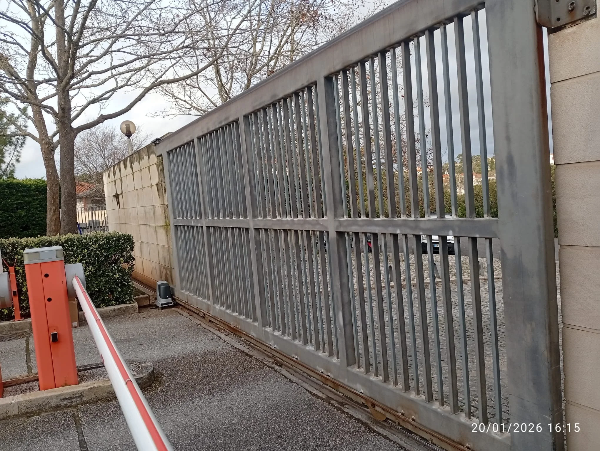 Imagem de uma cerca de grade metálica em uma entrada, ao lado de uma parede de pedra e uma parede de concreto, com árvores sem folhas ao fundo e uma luz de poste. Há uma cancela automática preta na base da grade e uma barreira de controle de acesso laranja e branca na parte inferior esquerda da imagem. O céu está nublado.