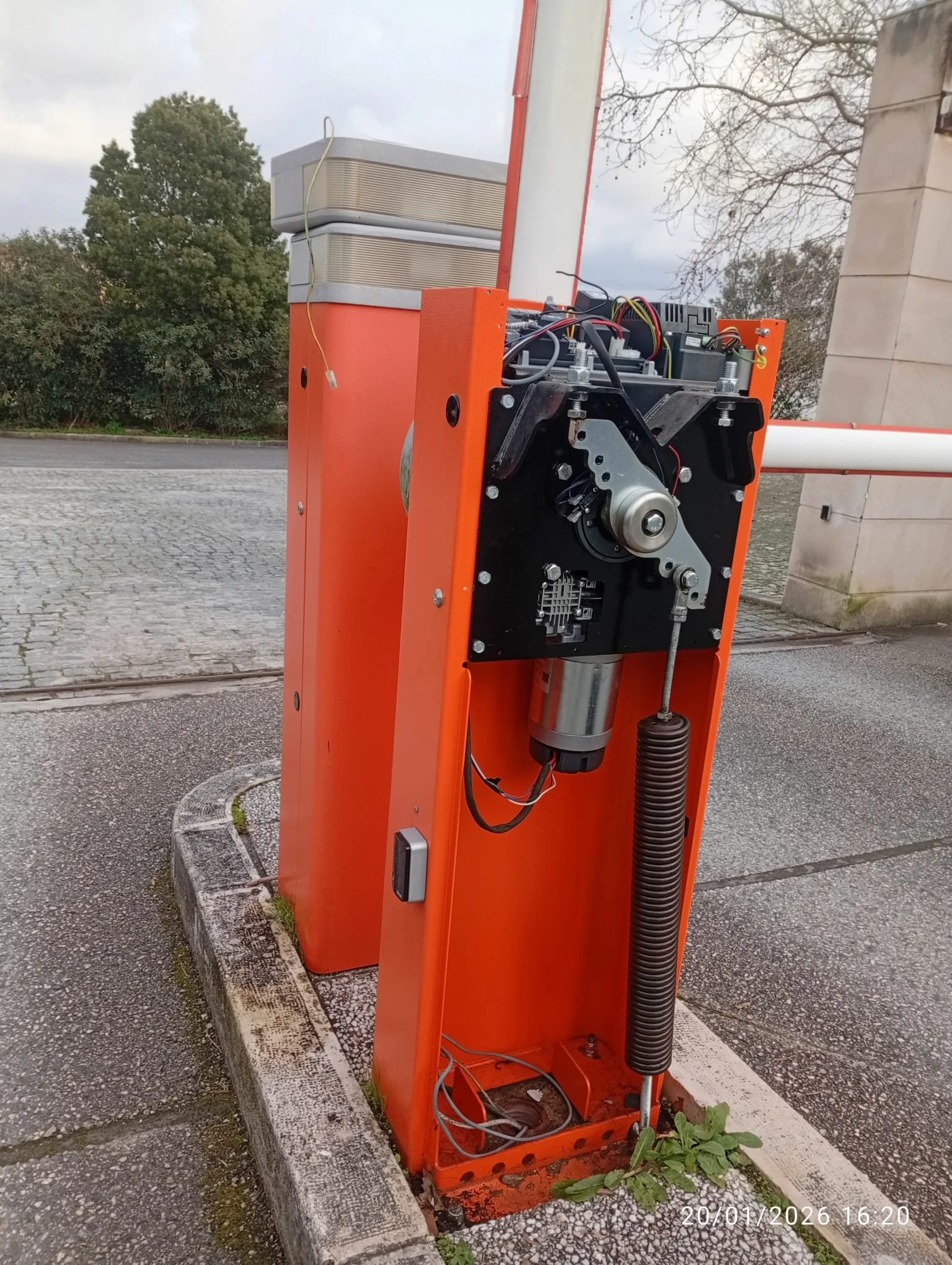 Imagem de um dispositivo de controle de acesso, possivelmente um portão automático, sem a carcaça de proteção, mostrando componentes eletrônicos internos, fixado em uma guia de estacionamento.