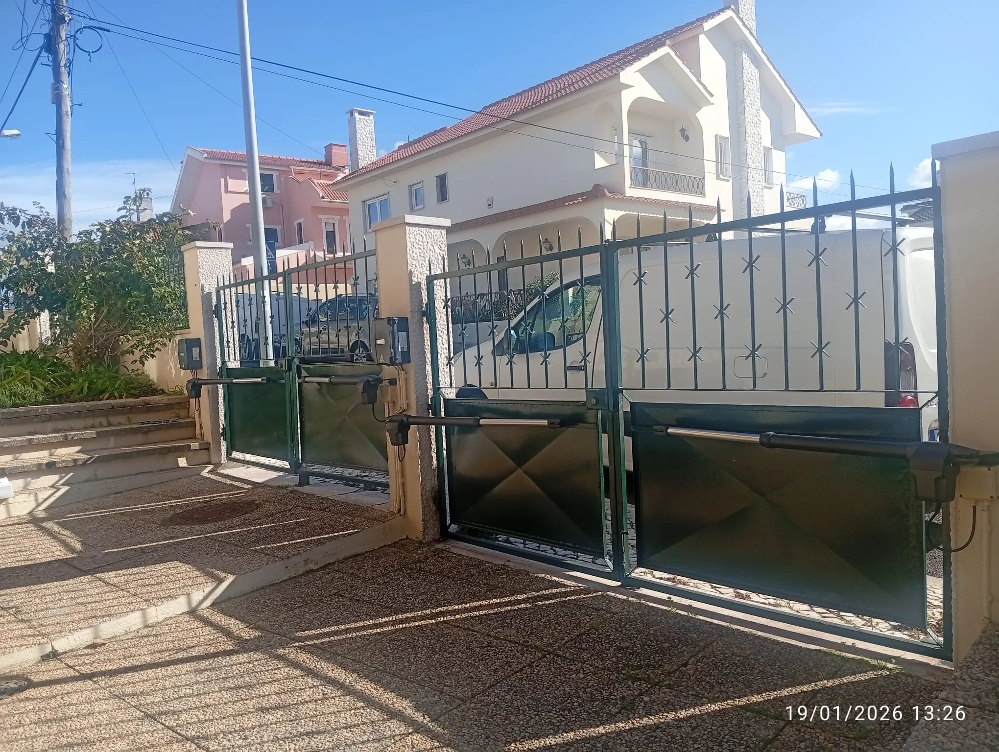 Vista de uma casa de dois andares com telhado de telhas vermelhas, fachada branca, cercada por uma cerca de metal, com carros estacionados na garagem e um jardim com plantas ao lado. É dia ensolarado com céu azul.
