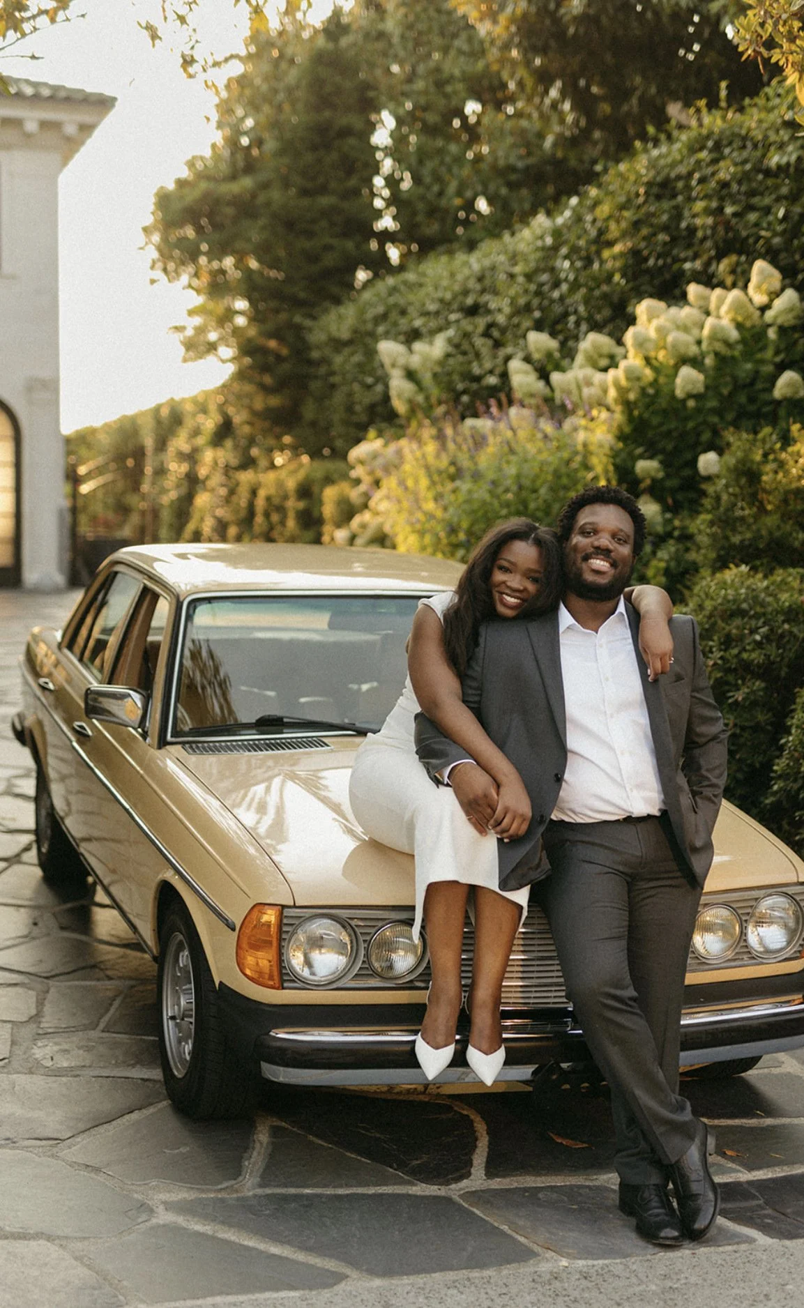 A happy couple sitting on the hood of a vintage beige car parked in a driveway, surrounded by lush green bushes and trees, with the woman hugging the man from the side.