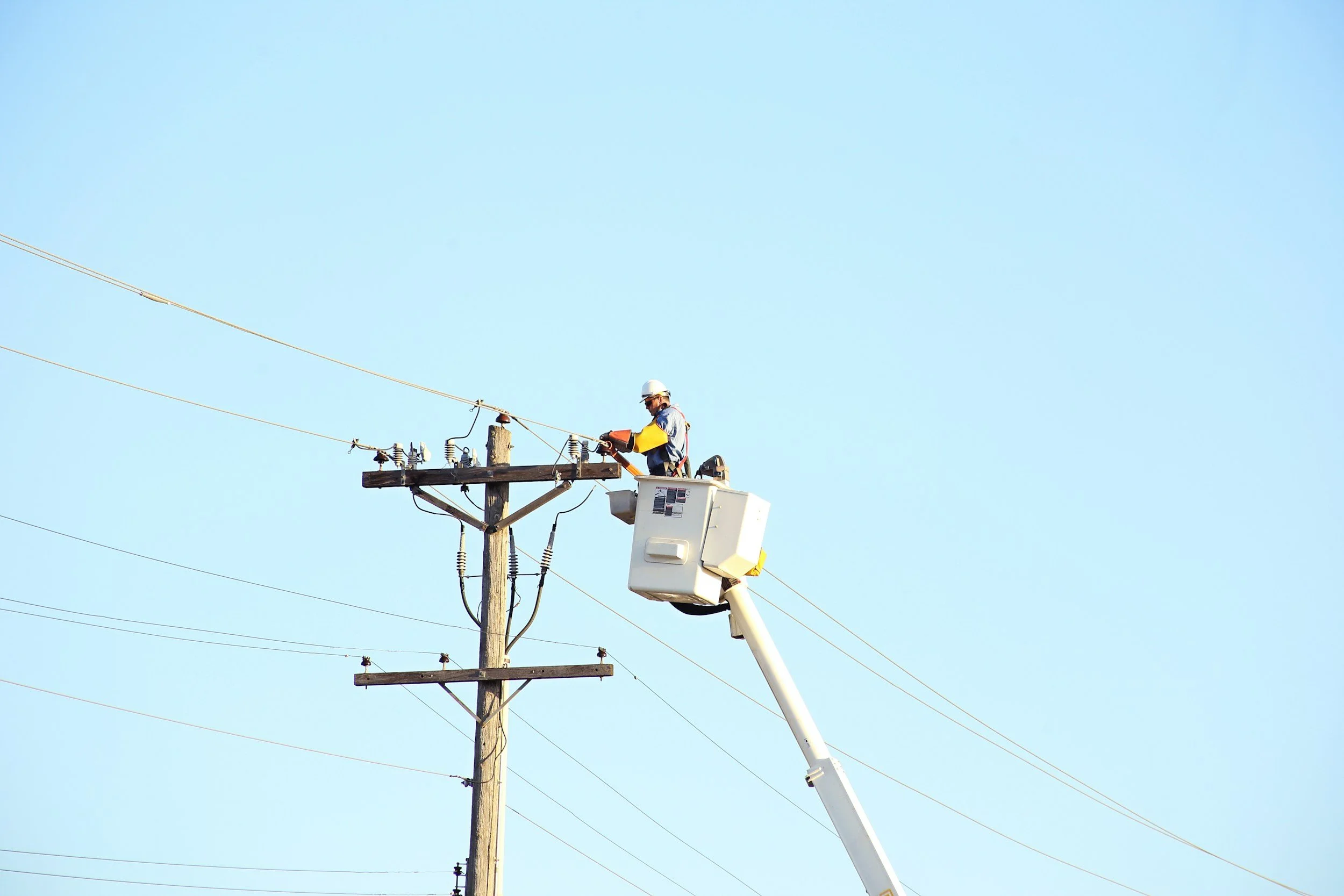 A utility worker wearing a helmet and safety gear is working on electrical lines from an elevated bucket lift, on a clear day with a bright blue sky.